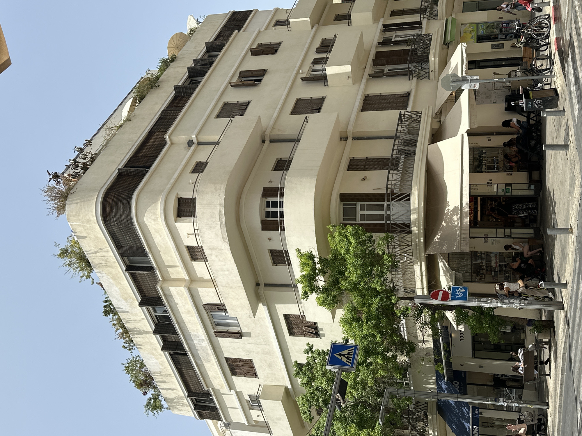The image shows a multi-story building in Tel Aviv, Israel, displaying a modern architectural style with rounded corners and light-colored facade. Balconies extend from various floors, some with shutters and some with plants. Street-level activity includes people sitting and walking on the sidewalk and there are parked bicycles. Trees and street signs are visible in the foreground.