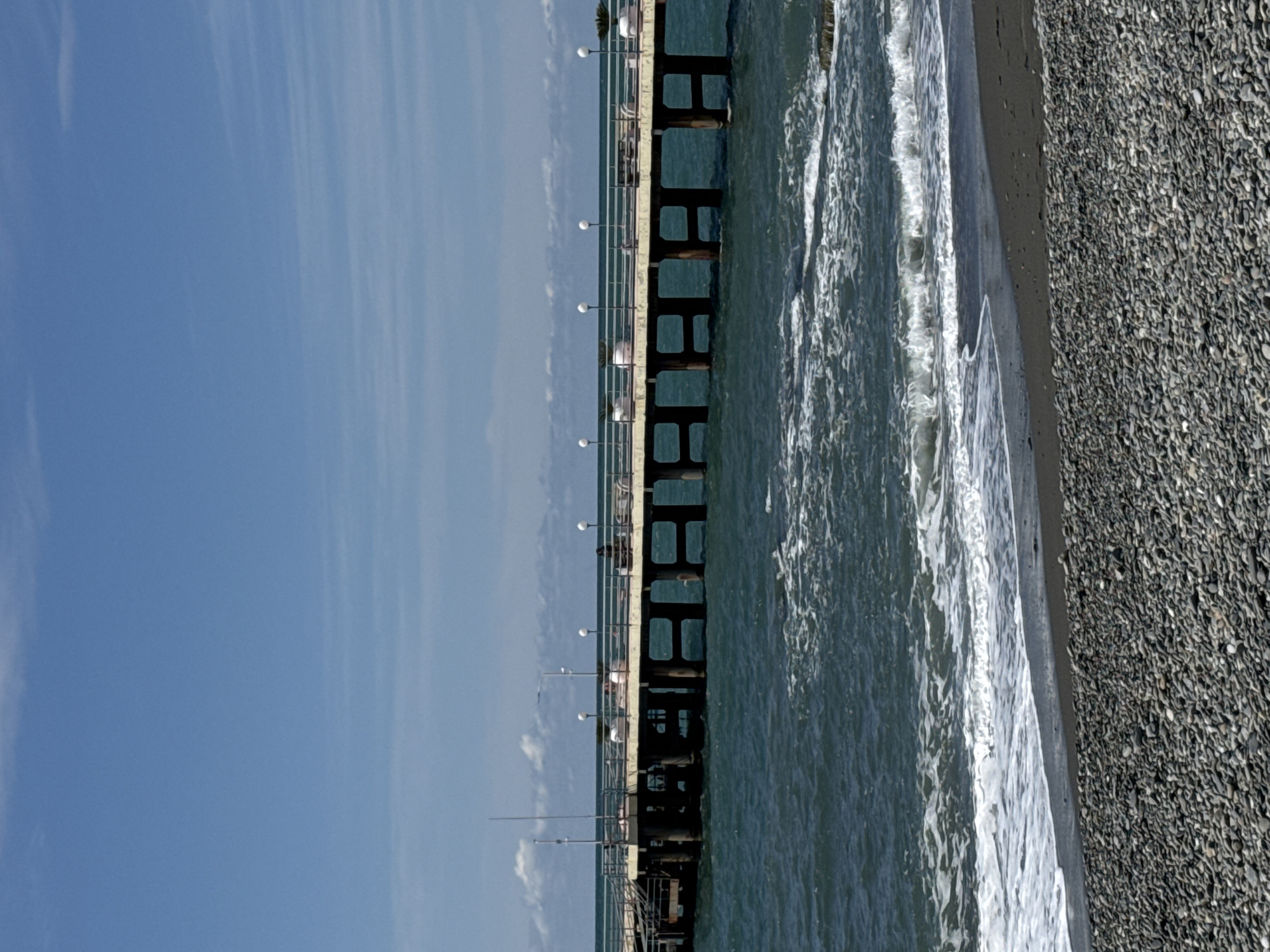 The image depicts a pier stretching out into the Black Sea, with a pebble beach along the shore. The sky is clear and blue. The pier is constructed of concrete pillars and has railings along its length. There are evenly spaced streetlights along the pier. The water is visible between the pier supports. Waves are breaking on the beach. The location is the city of Sochi, Russia.