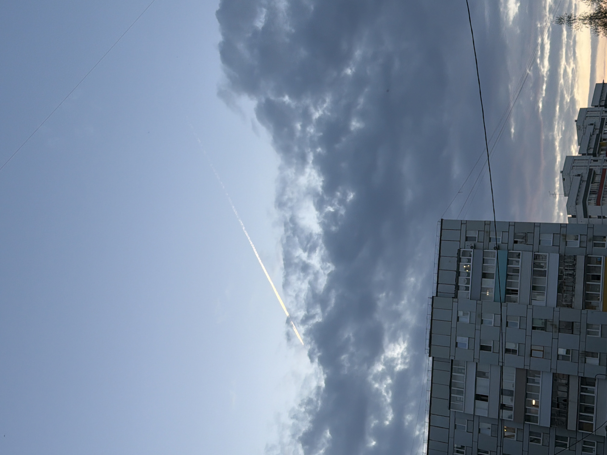 The image shows a view of the sky in Chaikovsky, Russia. A white contrail stretches across a blue sky with a line of dark gray clouds on the right. A building with many windows is visible on the right side of the image. There are also some utility wires crossing the sky.