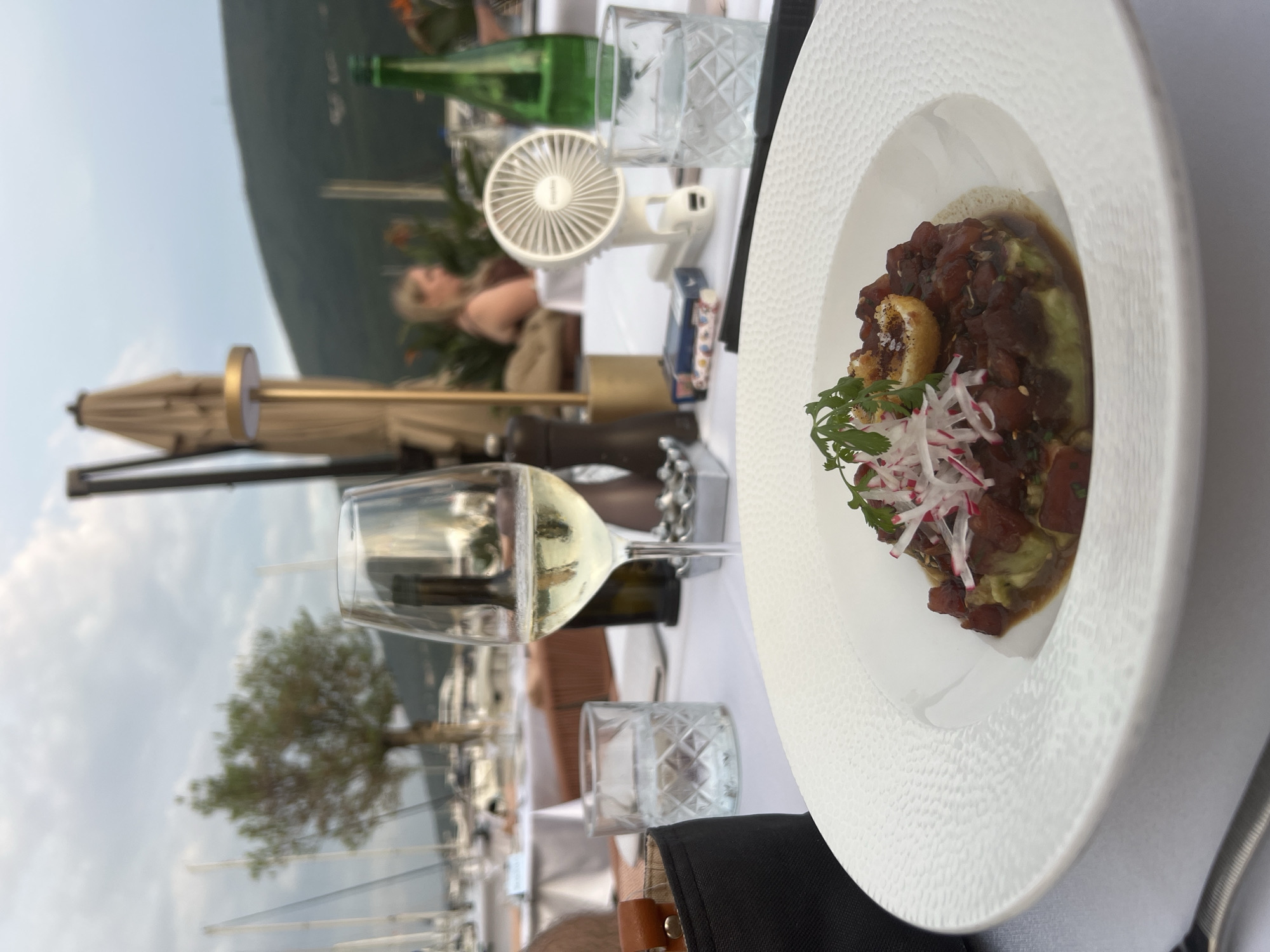 An overhead, close-up shot captures a white plate of tuna tartare on a white tablecloth at an outdoor dining establishment. The tartare is composed of cubed red tuna, avocado, and radish slivers, garnished with a sprig of cilantro and a round, fried garnish. To the left of the plate, a wine glass is filled with white wine, and behind it, a glass of water. A black clutch bag is positioned to the left of the wine glass. In the background, a green bottle of water sits on the table, next to a small white portable fan and a pack of cigarettes. Further back, white tablecloths adorn other tables, and a blurry figure of a woman is visible. Beyond the tables, a harbor with sailboats and a tree can be seen, with a tree-covered hillside in the distance. The scene is set in Đenovići, Montenegro.