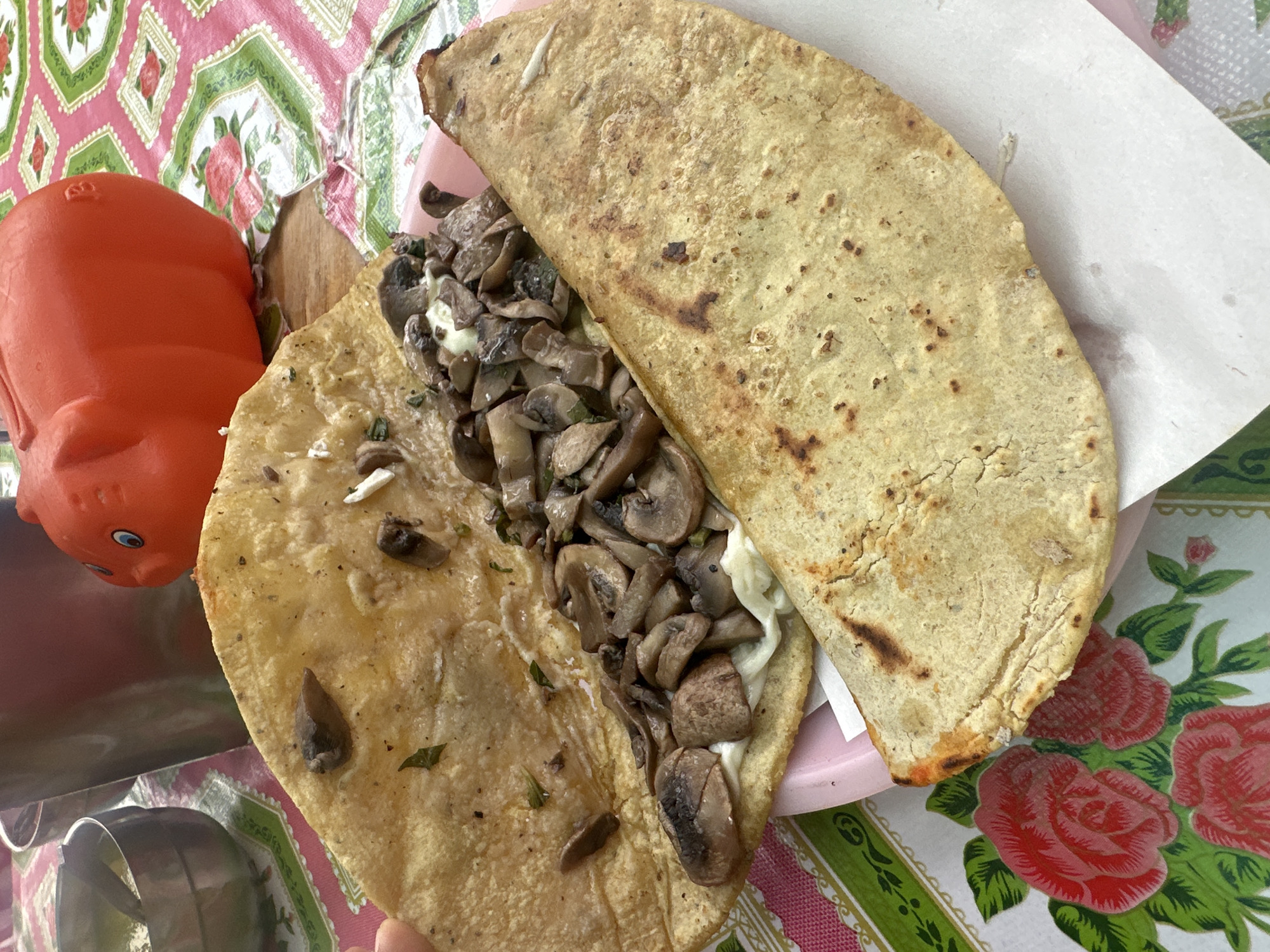 In San Miguel Zinacantepec, Mexico, a pink floral tablecloth background supports a pink plate holding a large quesadilla filled with mushrooms and white cheese. Next to the plate is an orange, pig-shaped piggy bank. A knife is partly visible on the bottom left corner. The quesadilla is cut in half, exposing the abundant mushroom filling.