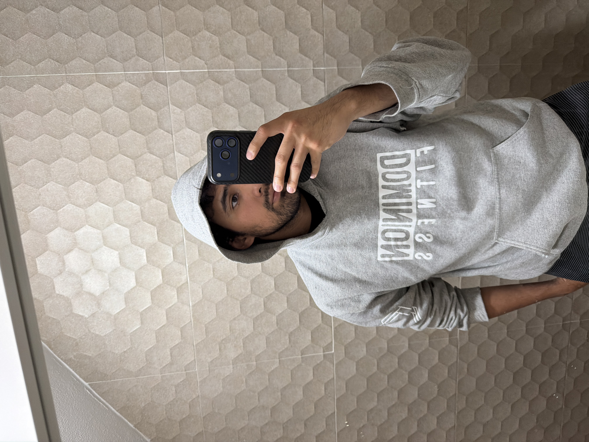 A person is taking a selfie in front of a wall covered with beige hexagonal tiles. The person is wearing a grey hooded sweatshirt with the words "DOMINION FITNESS" printed on the chest in a mirrored image. The hoodie is pulled up over their head. They are holding a dark-colored smartphone in their right hand, obscuring part of their face. The wall appears to be a bathroom or tiled surface of some sort. The city of Cuautla, Mexico is not discernible from the image.