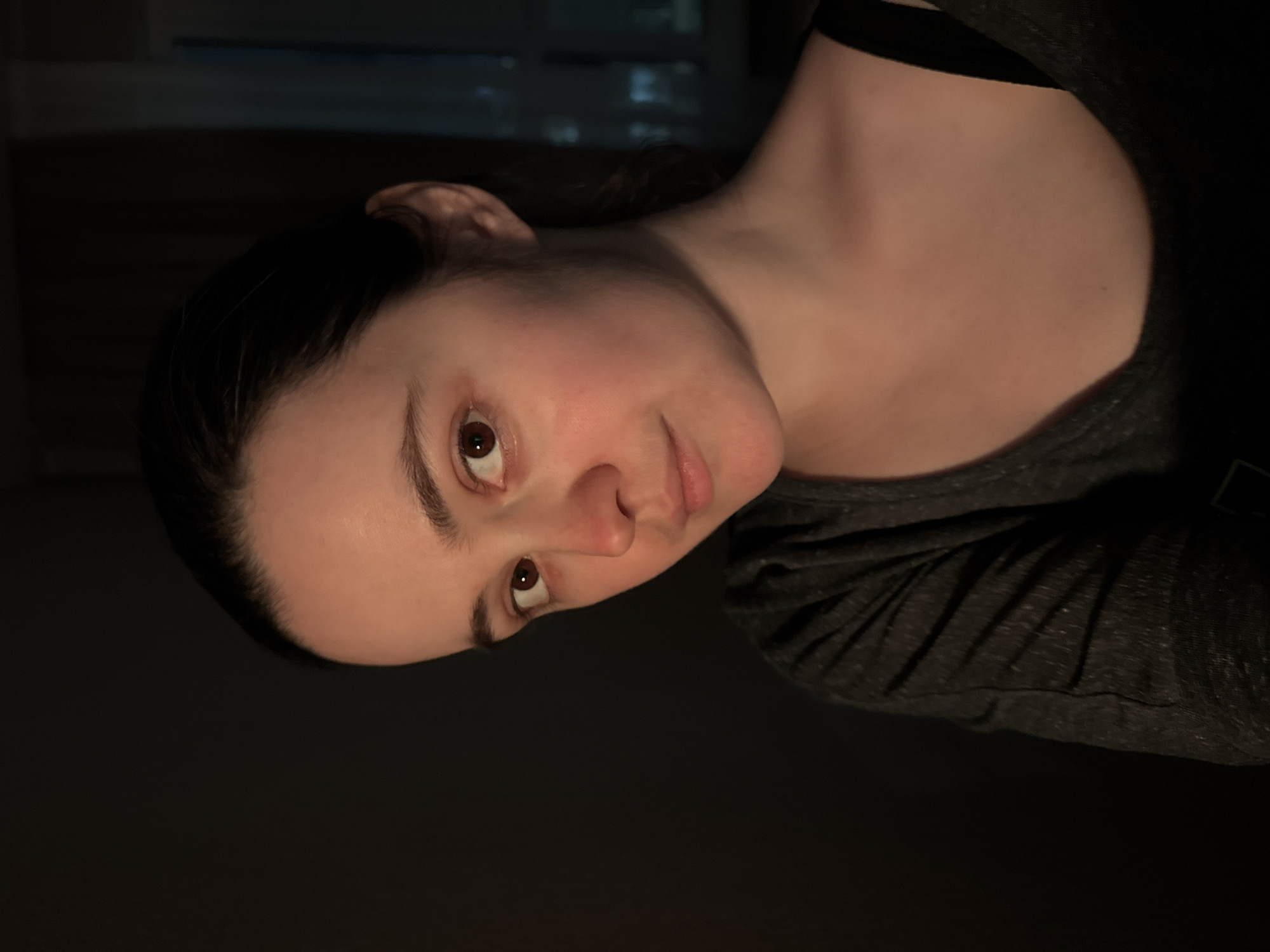 A woman with dark hair and light skin is shown in a portrait oriented image. Her head is tilted to her right. She is wearing a dark gray shirt with a black tank top strap visible at the top. The background is dark. It is not possible to ascertain the location of the scene, including whether it is in Izhevsk, Russia, from the image provided.