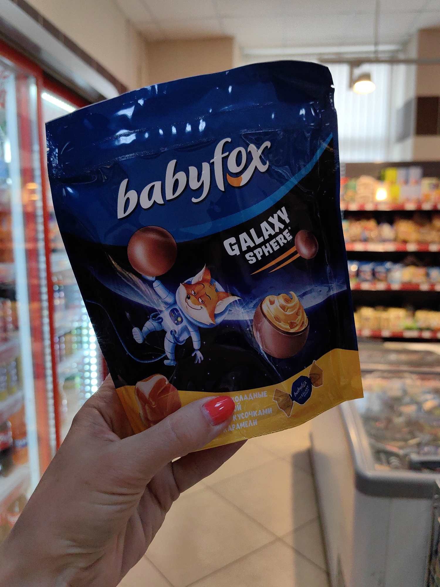A hand with bright red nail polish holds a sealed, dark blue and yellow pouch of "babyfox GALAXY SPHERE" chocolate candies in front of a blurred background of grocery store shelves filled with products. The pouch features an illustration of a cartoon fox in an astronaut suit floating in space with chocolate spheres and a depiction of a chocolate sphere with caramel filling. Text in Cyrillic on the lower part of the pouch reads "ШОКОЛАДНЫЕ В КУСОЧКАМИ КАРАМЕЛЕЙ" which translates to "CHOCOLATE WITH CARAMEL PIECES". The background suggests a retail environment with brightly lit refrigerators and merchandise displays. The lighting is ambient store lighting. The image is taken from a slightly low angle, looking up at the product and the hand.