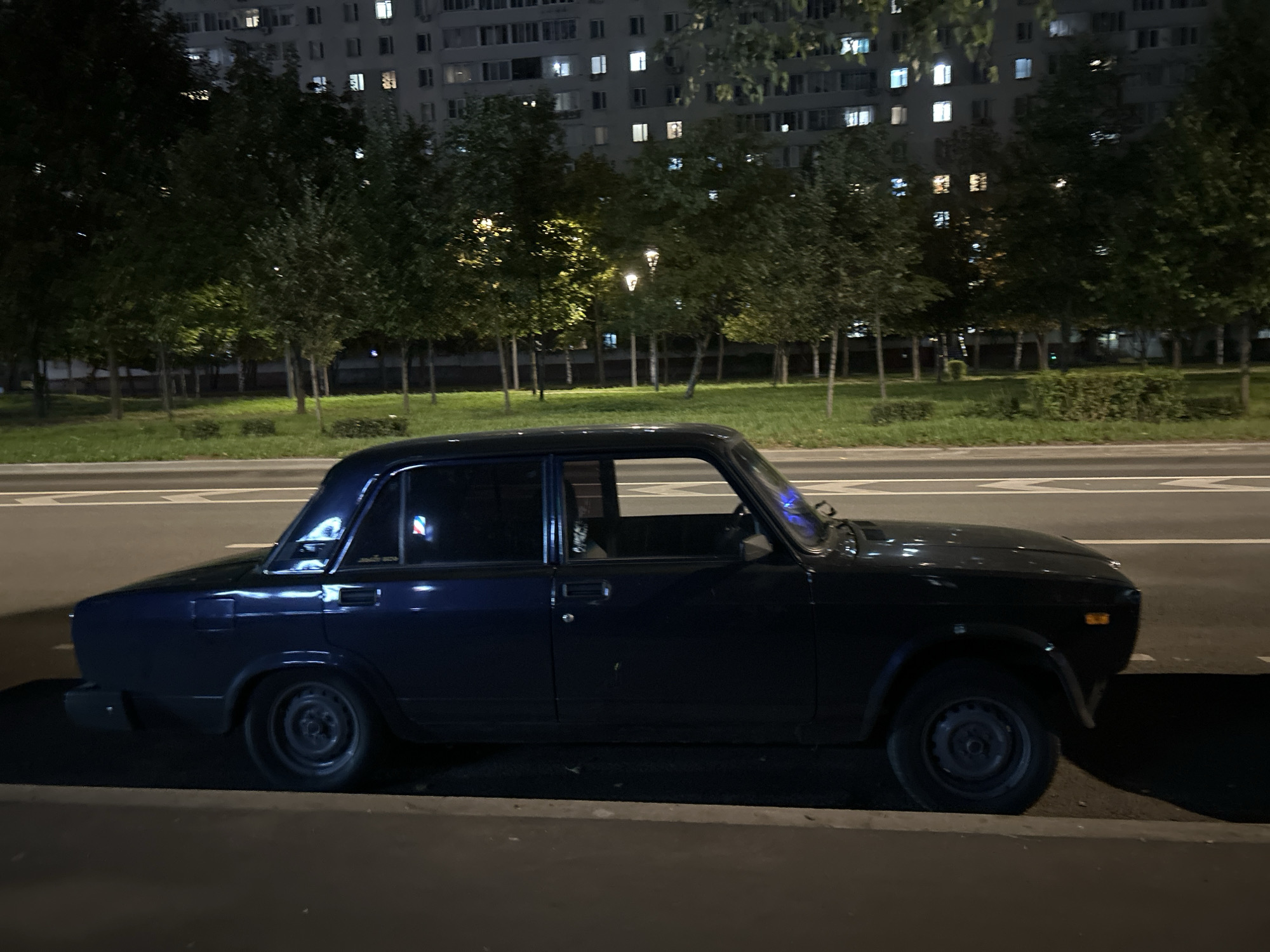 A dark blue sedan is parked on the side of a road at night. The car is positioned parallel to a curb, with its tires on the asphalt. Behind the car, there is a strip of grass and then a row of trees. In the background, a large residential building with numerous lit windows is visible. The street appears to be a multi-lane road with white markings. The overall scene is characteristic of a residential area in Moscow, Russia, at night, with streetlights and building lights illuminating the surroundings.