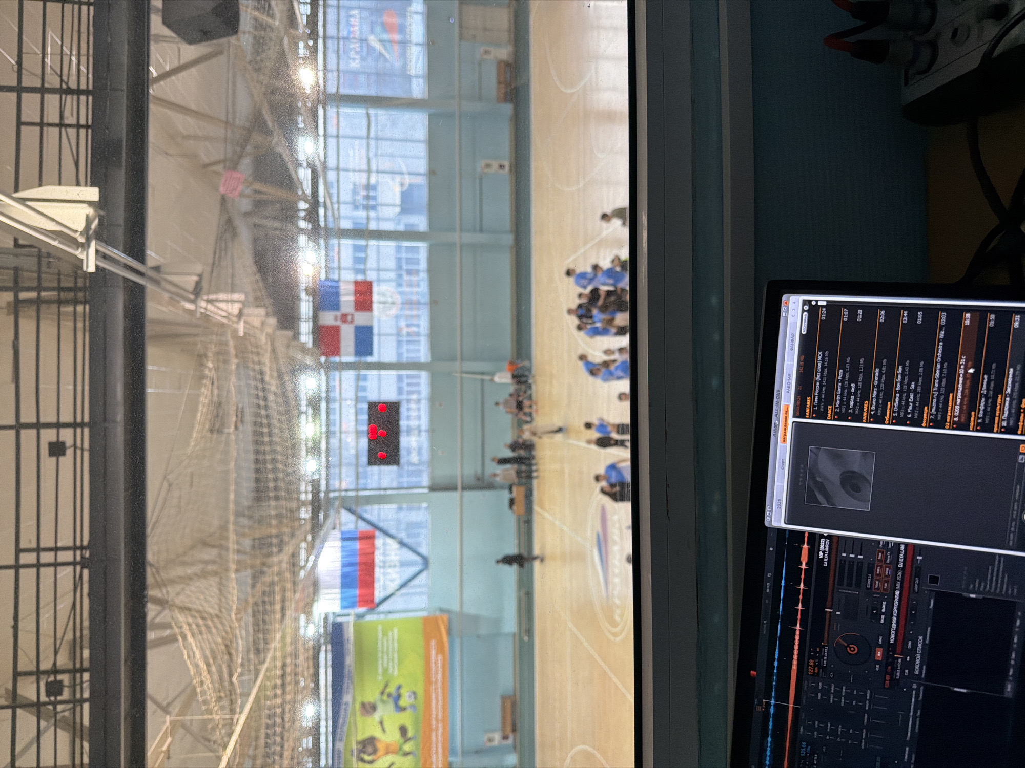 The image shows a view from inside a room, looking out through a window at an indoor sports court in Kondratovo, Russia. The sports court features basketball hoops, court markings, and a digital scoreboard. A gathering of people, possibly a team or group, is visible on the court. There are flags of the Dominican Republic and Russia hanging in the gym. In the foreground, inside the room, a computer screen displays audio editing software with various audio files listed.