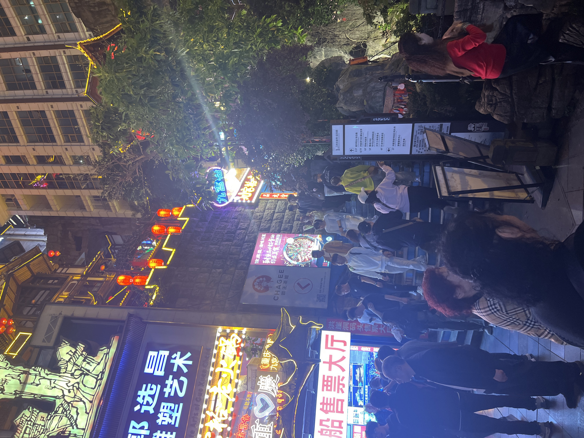 The image shows an evening scene in Yuzhong District, Chongqing, China. A crowd of people walks on a sloped stone walkway illuminated by various neon signs in Chinese characters and strings of red lanterns. A multi-story building with traditional Chinese architectural elements is visible to the left. Several commercial signs, including those for "CHAGEE" and "洪崖洞码头售票大厅" (Hongyadong Wharf Ticket Hall), are displayed. A directory sign indicates various attractions on different floors. People are walking, and some are looking at the illuminated signs and attractions.