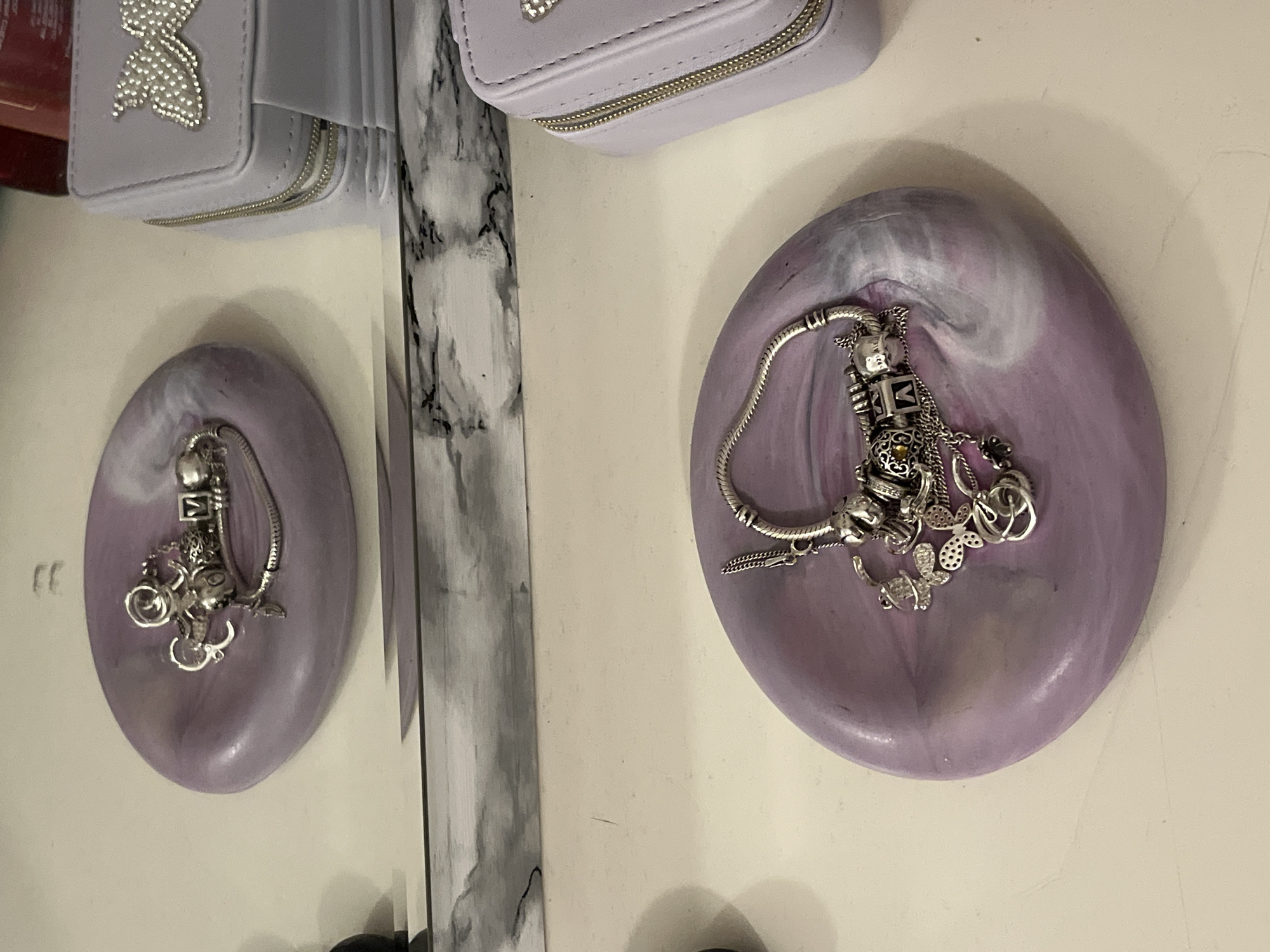 The image displays a still life arrangement of personal items, predominantly jewelry, set on a light-colored surface in an indoor environment. The central subject is a light purple, oval-shaped trinket dish exhibiting a subtle white marbled pattern. Resting within this dish is a silver-colored charm bracelet, laden with various intricate charms. Notable charms include block letters (one appearing to be an 'A' and another a 'B'), a small metallic figure resembling a robot, butterfly designs, and other decorative elements, some featuring tiny gemstones. A portion of a fine chain also dangles from the bracelet.
To the upper left, a small, light purple rectangular case with a gold-colored zipper is visible. The lid of this case is adorned with a silver-colored butterfly embellishment crafted from small studs or rhinestones. A vertical mirror positioned towards the left of the frame reflects both the purple dish with its bracelet and a segment of the purple case, creating a mirrored composition of these items.
The background includes a vertical element with a black and white marble-like pattern, likely a decorative stand or part of the mirror's frame. The underlying surface holding these objects is uniformly off-white or light beige. A small, dark red object is partially glimpsed in the far upper-left corner of the image. The scene is illuminated by soft, diffused lighting, characteristic of an indoor setting. There is no discernible activity occurring, nor is there any clear, readable text visible on any of the depicted objects.