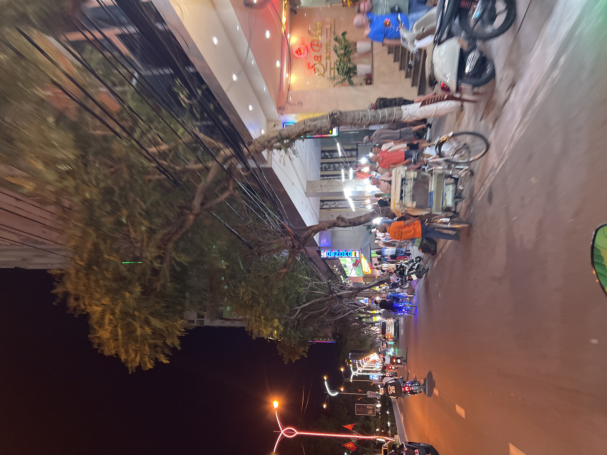 The image depicts a bustling street scene at night in Nha Trang, Vietnam. The camera appears to be positioned on the street, facing down a boulevard. Streetlights illuminate the multi-lane road where motorbikes and vehicles are actively moving. A sidewalk runs alongside the road, crowded with pedestrians and parked motorbikes. There is some business activity as several people can be seen eating and socializing. Power lines crisscross the sky above the trees lining the sidewalk. Storefronts with bright lights and signs are also visible.