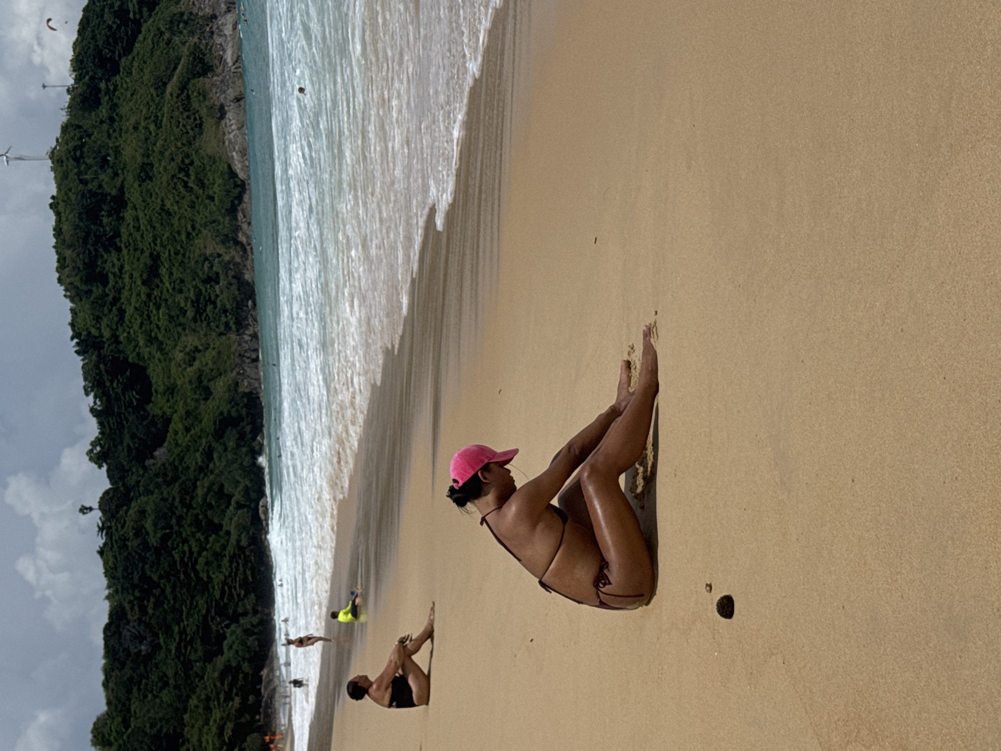 The image depicts a beach scene in Rawai, Thailand. In the foreground, a woman wearing a brown bikini and a pink baseball cap is sitting on the sand with her legs extended. Her body is tanned, and she appears to be resting. Further back on the beach, another woman in a black bikini is also sitting. In the middle ground, several people are visible near the water's edge, including a person in a yellow shirt sitting down. The ocean is a vibrant blue with white waves rolling onto the shore. Behind the beach, a densely forested hillside rises, with a few structures, including wind turbines, visible at the top. The sky is overcast with some patches of blue. The sand is light brown and appears to be slightly damp near the water. There are a few small shells and pebbles scattered on the sand.