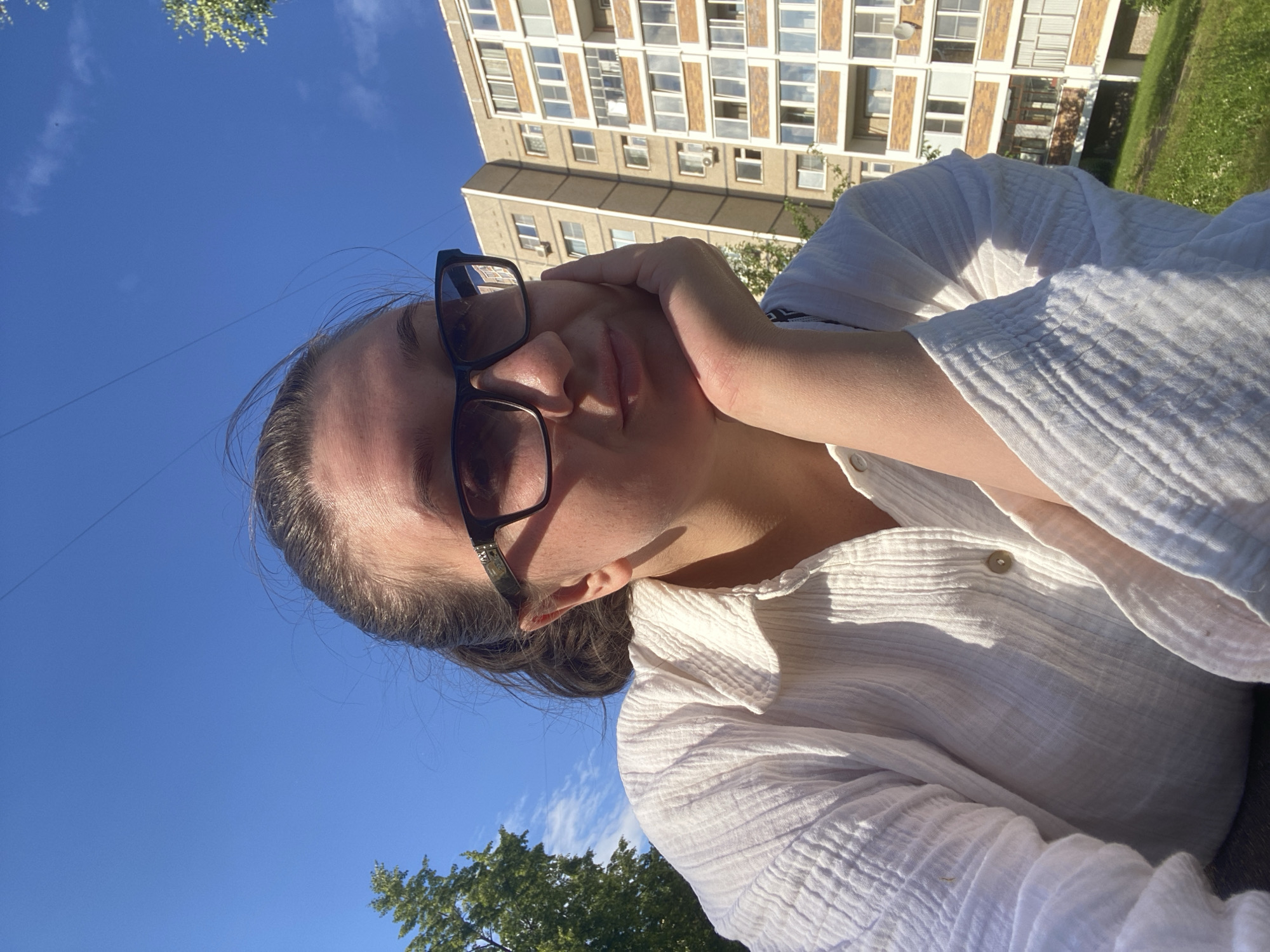 A young woman with her hair in a ponytail, wearing glasses and a white, textured shirt, rests her chin on her hand. She is outdoors, with a bright blue sky overhead. In the background, a tall, multi-story apartment building with numerous windows stands against the sky. The building appears to be concrete and has several balconies. Green grass and trees are visible at the base of the building and on a slight incline to the left. The scene suggests a daytime setting, possibly in a residential area of Chaikovsky, Russia.