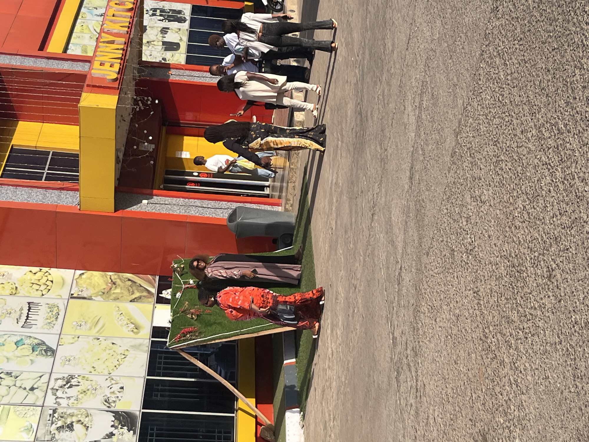 The image shows the exterior of Jenny Kitchen, a restaurant in Gura Top, Nigeria. The building is painted in red, yellow, and orange. Several people are present, including groups standing and walking outside the restaurant. The area in front of the restaurant is paved.