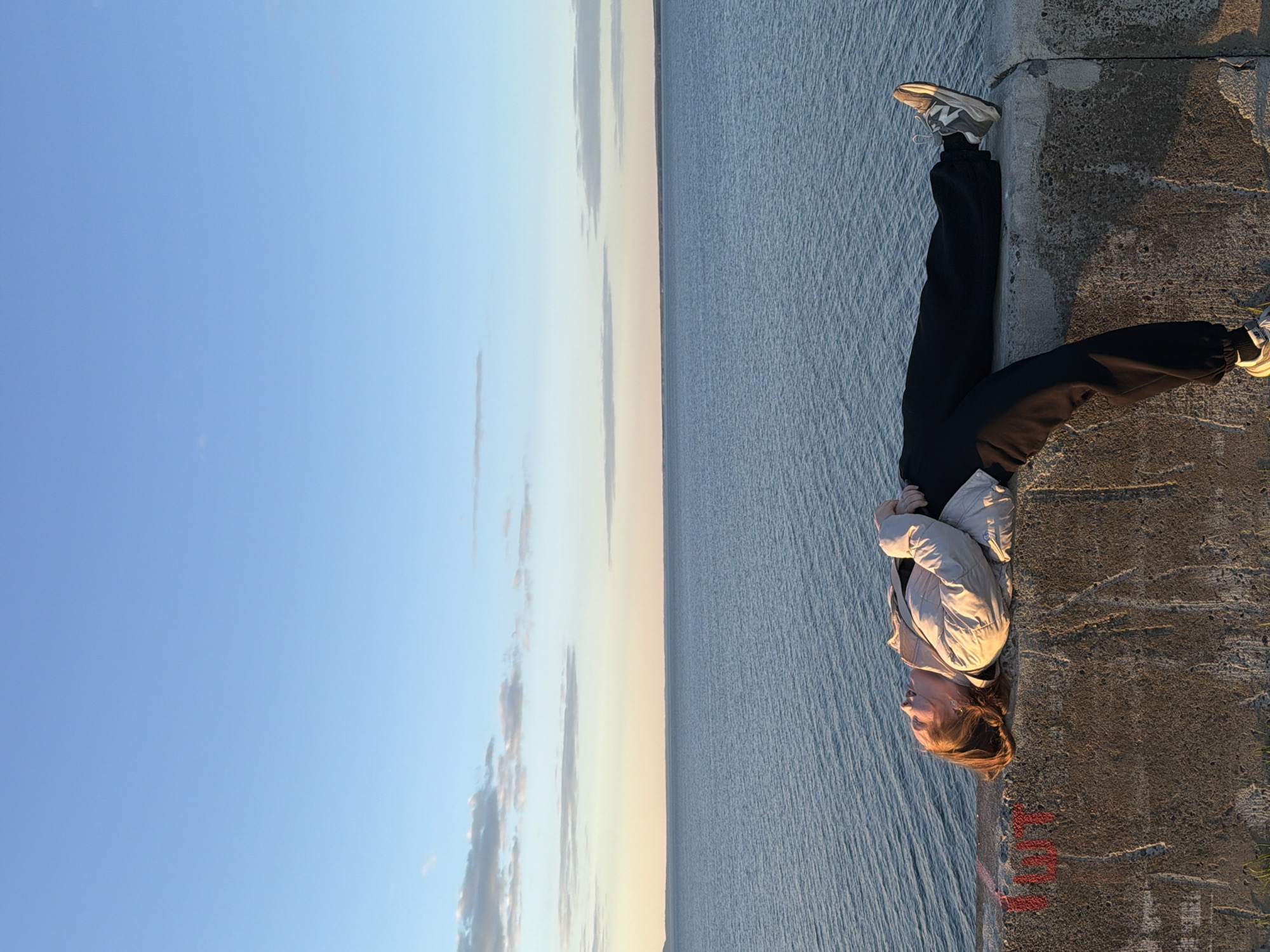 A person wearing black pants, a light-colored jacket, and sneakers stands on a concrete structure, with water to their left and a blue sky with light clouds behind the water. The location may be in Chaikovsky, Russia.