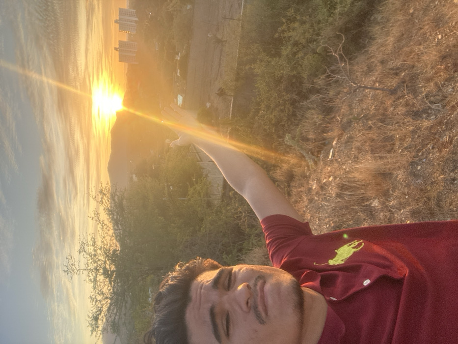The image captures a male individual positioned in the foreground, approximately from the chest up, against a sunset or sunrise landscape in Juan Griego, Venezuela. The subject, with dark hair, a mustache, and a subtle goatee, wears a red polo shirt adorned with a light green polo player emblem on the left chest. He extends his right arm, palm open, towards the bright, golden-orange sun, which is prominent in the upper right portion of the frame and emits distinct light rays. His facial expression displays a slight pout or smirk, with his eyes partially narrowed. The immediate foreground features dry, yellowish-brown grass and sparse vegetation. In the mid-ground, a range of dark hills or mountains is silhouetted. The distant background on the right reveals two multi-story buildings, consistent with an urban or coastal setting, along with additional vegetation and a flatter area of terrain. The sky exhibits a gradient of light blue, orange, and yellow hues, with scattered, wispy clouds.
