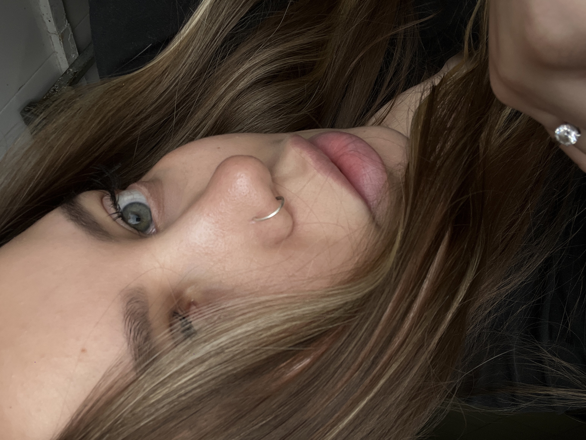A close-up, low-angle view of a fair-skinned woman with light brown hair. She has a silver nose ring on her left nostril and a stud earring. Her eye is light green. The background appears dark, possibly clothing. The image contains no discernible elements to indicate location in Novopokrovskaya, Russia.