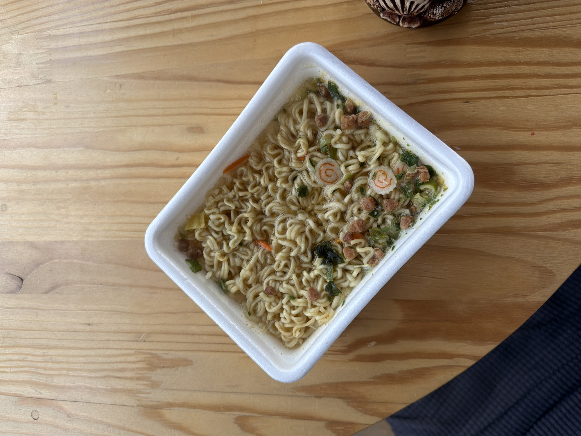A top-down, medium shot of a white, square container filled with cooked ramen noodles is centered on a light-brown wooden surface. The noodles are interspersed with small pieces of green vegetables, orange shredded carrots, and sliced pink and white fish cakes. A dark object with visible texture is present in the bottom-right corner of the image. Another, unidentified object sits in the top-right corner. The location is Unknown, Unknown.
