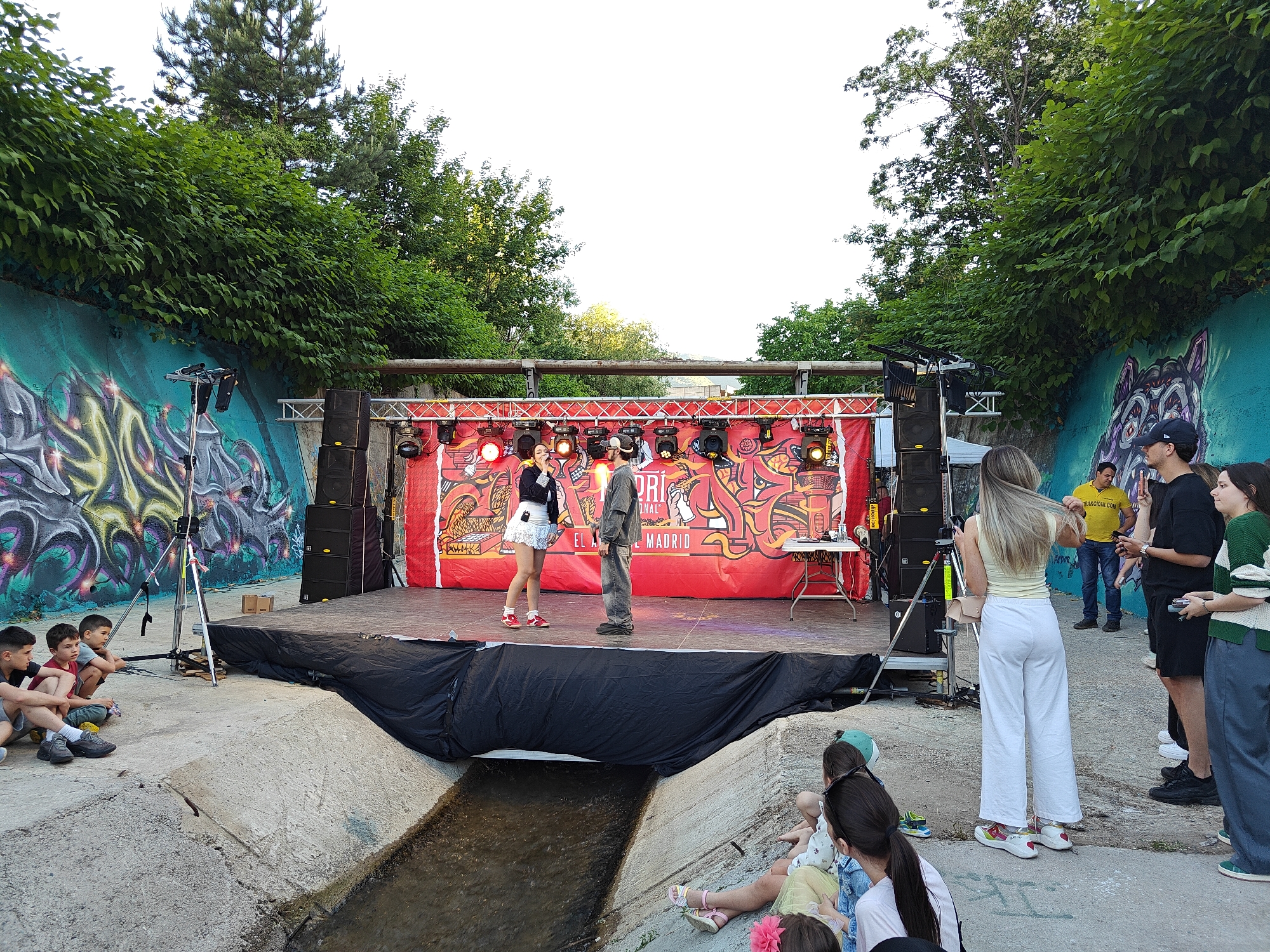The scene depicts an outdoor performance in what appears to be a converted concrete drainage channel in Sofia, Bulgaria. A raised stage is set up with a red backdrop featuring graphic art and the word "MADRID." Two individuals, a woman in a white skirt and a man in a grey outfit, are on stage, possibly performing. The stage is equipped with speakers and lighting. A small group of children are seated on the ground to the left of the stage, watching the performance. To the right, a group of adults are standing and observing. The background features graffiti-covered walls and green trees and bushes, creating an urban yet natural environment. A shallow stream of water flows in front of the stage.