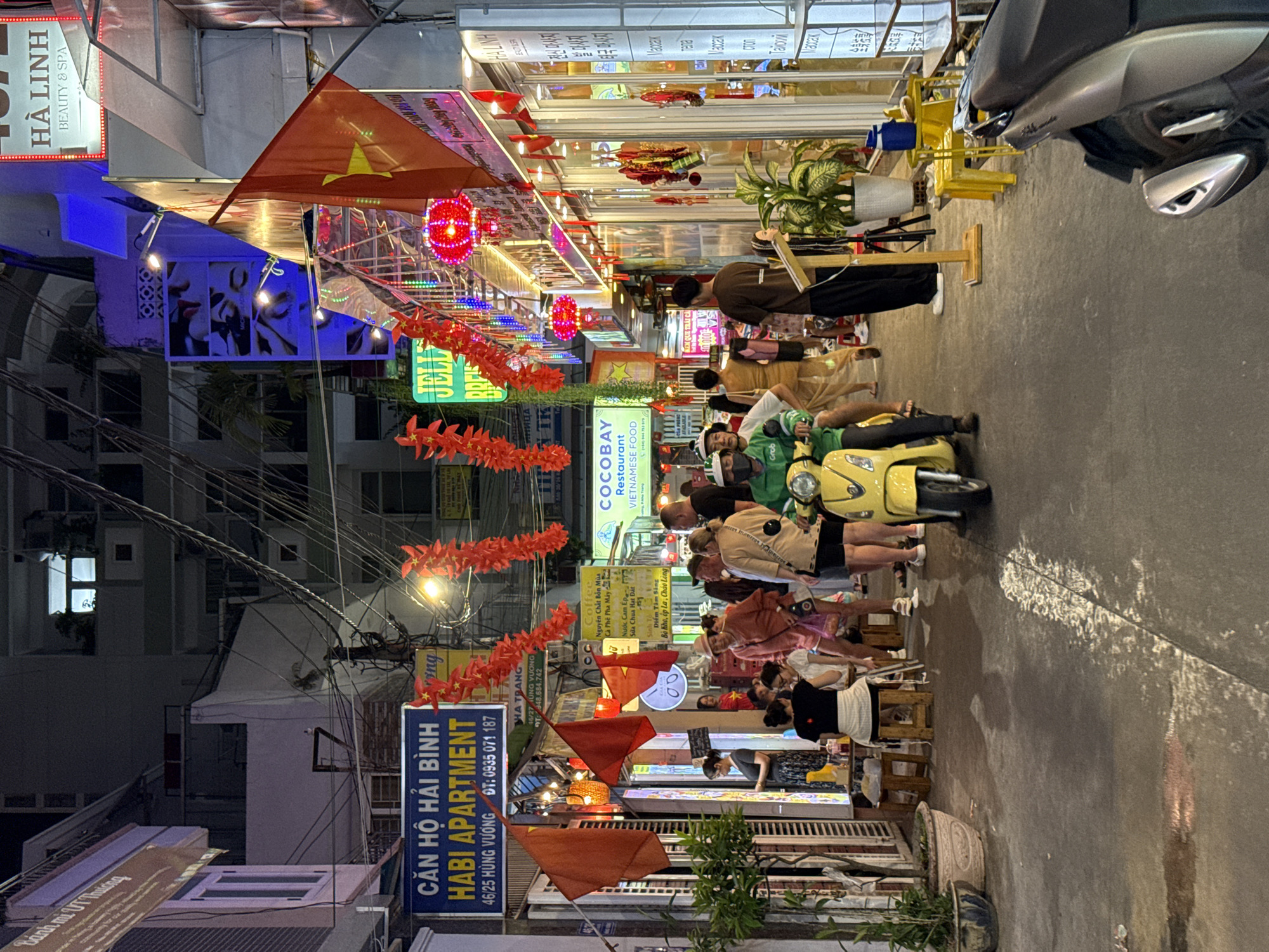 The image shows a narrow street scene in Nha Trang Ward, Vietnam, at night. The street is lined with small restaurants and businesses, indicated by illuminated signs in both Vietnamese and English. Red lanterns and flags adorn the street. People are seated on small stools, likely dining at the restaurants. A yellow motorbike is parked on the side. Overhead power lines are visible. The image is taken from a slightly elevated position.