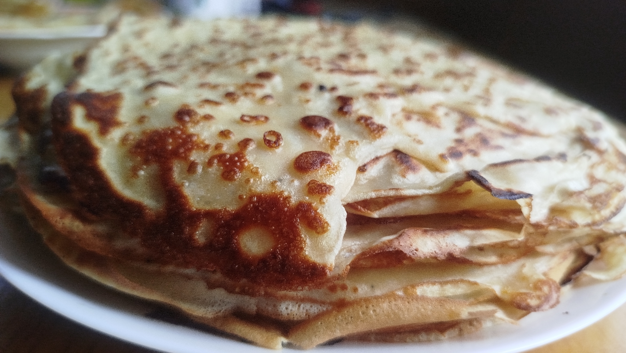 A close-up, slightly blurred photograph shows a stack of golden-brown crepes on a white plate. The crepes are thin and have slightly crispy edges with some darker brown, almost burnt, spots. The texture of the crepes is visible, with small bubbles and irregular surfaces. The lighting casts a soft glow on the food, highlighting its texture and color. The background is dark and out of focus, suggesting an indoor setting. The image does not contain any identifiable landmarks or features that would place it specifically in Kostiantynivka, Ukraine, or any other city. There are no people, actions, or interactions depicted.