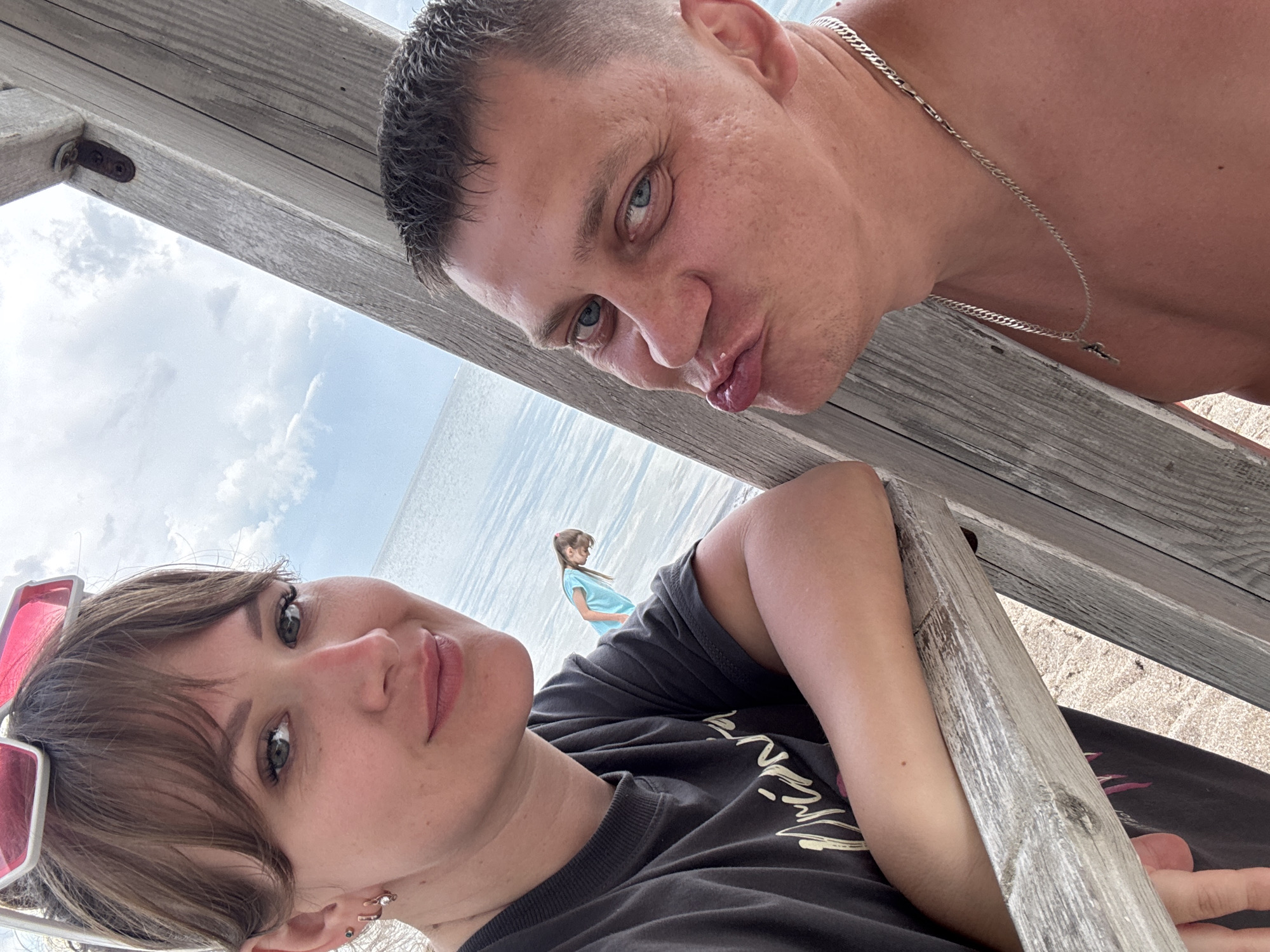 A low-angle selfie shows a man and a woman, seemingly under a wooden structure, likely at a beach based on the beach sand and sea in the background. A girl stands alone in the distance by the water.
The man, positioned to the upper right, has short, dark hair, light skin, blue eyes, and a silver necklace. He is making a pouty face.
The woman, in the lower left, has shoulder-length brown hair with bangs, light skin, and a dark-colored t-shirt. A pair of red sunglasses rest atop her head.
The location, inferred from the provided context, is Yevpatoriya, Ukraine, though this information is not evident from the image itself.