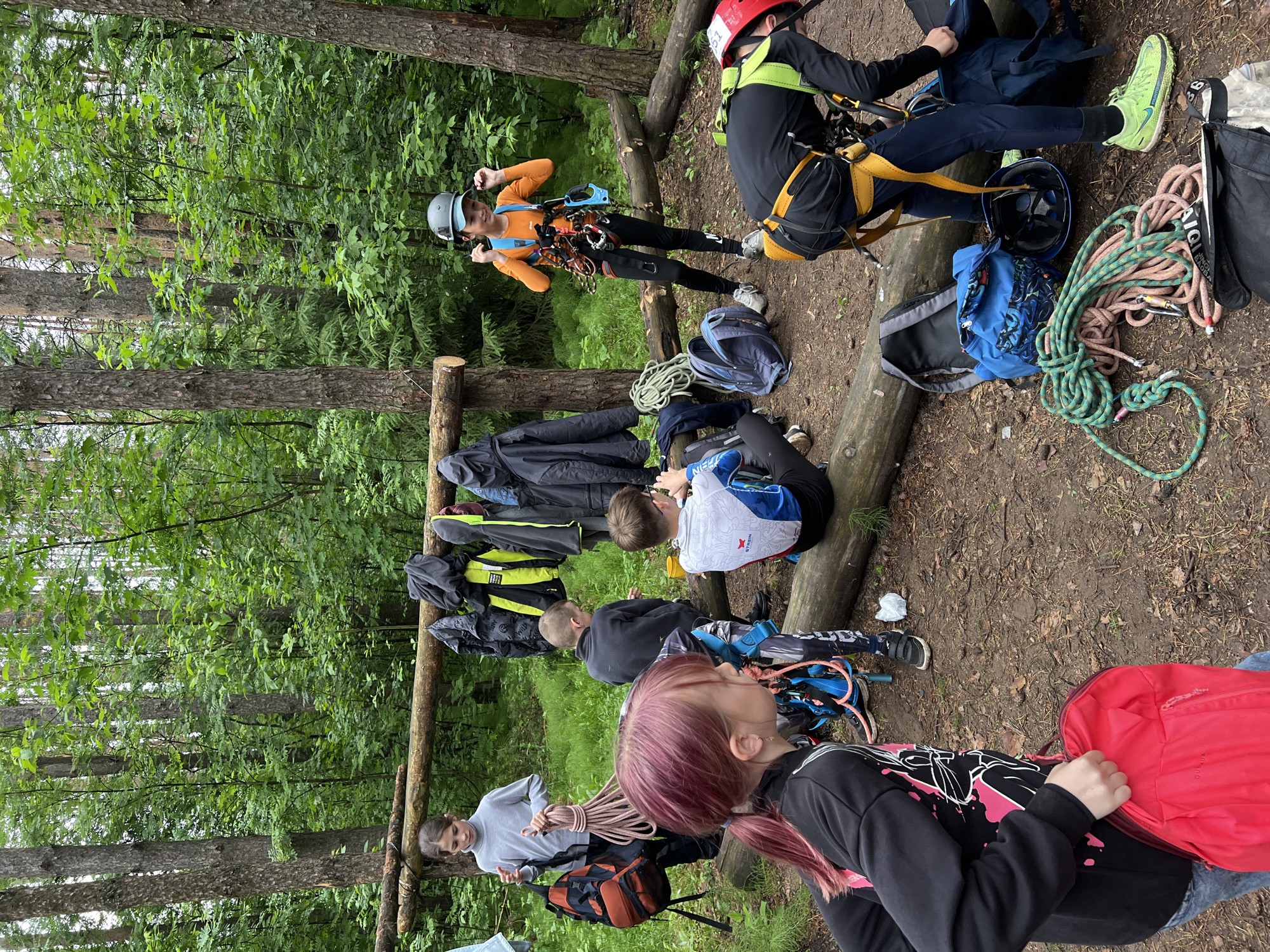 The image depicts a group of people, primarily young individuals, in a forest setting, likely preparing for or taking a break from an outdoor adventure activity such as climbing or a ropes course in Chaikovsky, Russia. The scene is set on a dirt ground surrounded by tall trees and dense green undergrowth, with several logs laid horizontally, possibly serving as seating or equipment racks.
Multiple individuals are visible, some actively engaged with gear, others resting. A person wearing an orange long-sleeve shirt, a gray helmet, and a climbing harness with carabiners attached stands confidently, flexing their arms. Another person, wearing a black long-sleeve top and a red helmet with the number "15" on it, is seated on the ground, seemingly adjusting their equipment. Other participants are seated on logs or the ground; one in a gray sweatshirt holds a coiled beige climbing rope, and another with pink hair wears a black sweatshirt. A person in a black and white patterned top is also seated on a log.
Various climbing and outdoor gear is present: coiled climbing ropes in green and beige, multiple backpacks (including a blue and a red one with "QUECHUA" visible), helmets, and harnesses. Jackets are seen hanging from a makeshift wooden rack. The ground appears slightly damp, and the natural light suggests it is daytime, possibly overcast or under a heavy tree canopy. A shoe with "SALOMON" visible on the side is also present. The overall mood appears focused and engaged in the outdoor activity.