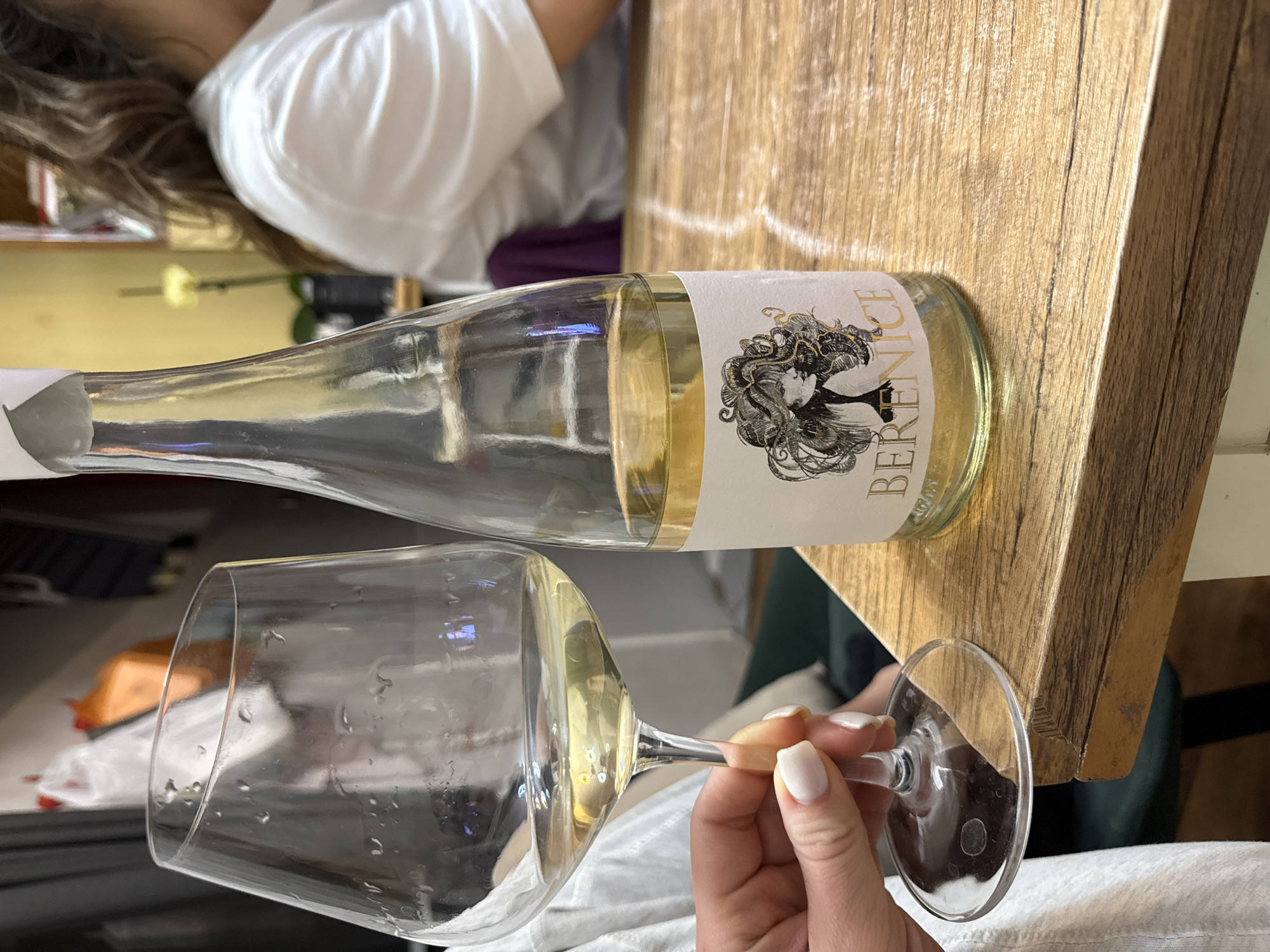 A photograph shows a partially filled wine glass held by a hand with white nail polish, next to a horizontal bottle of Berenice wine. The bottle has a white label with a black and white graphic of a woman's face and the name "BERENICE" printed on it. The wine appears to be white. The scene is set on a wooden surface, likely a table. The background is blurred but includes hints of a person in a white shirt. There is no information to identify the location as Plovdiv, Bulgaria, or any other specific city.