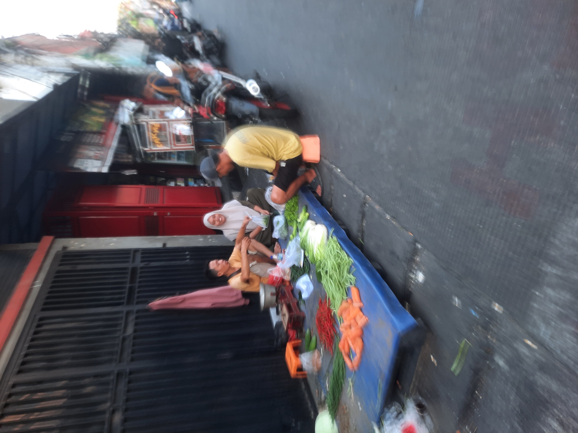 This is a medium shot, eye-level, outdoor photograph taken in the city of North Jakarta, Indonesia. The image captures a street scene with a group of people and a vegetable stall. The foreground features a blue tarp laid out on the ground, displaying a variety of fresh produce, including long beans, carrots, red chilies, and leafy greens. To the left, a black metal gate serves as a backdrop. In the center of the image, three individuals are present. A man in a yellow t-shirt and black shorts is seated on an orange stool, facing away from the camera. Beside him, a woman in a white hijab is also seated, holding a plastic bag. Another man, wearing a tan t-shirt and shorts, sits to the left of the woman. Behind the stall, a red building with closed doors is visible, with a dark awning extending over the street. Several motorcycles are parked to the right of the frame, indicating the urban setting. The overall impression is a glimpse into daily commerce and street life in this Indonesian city.