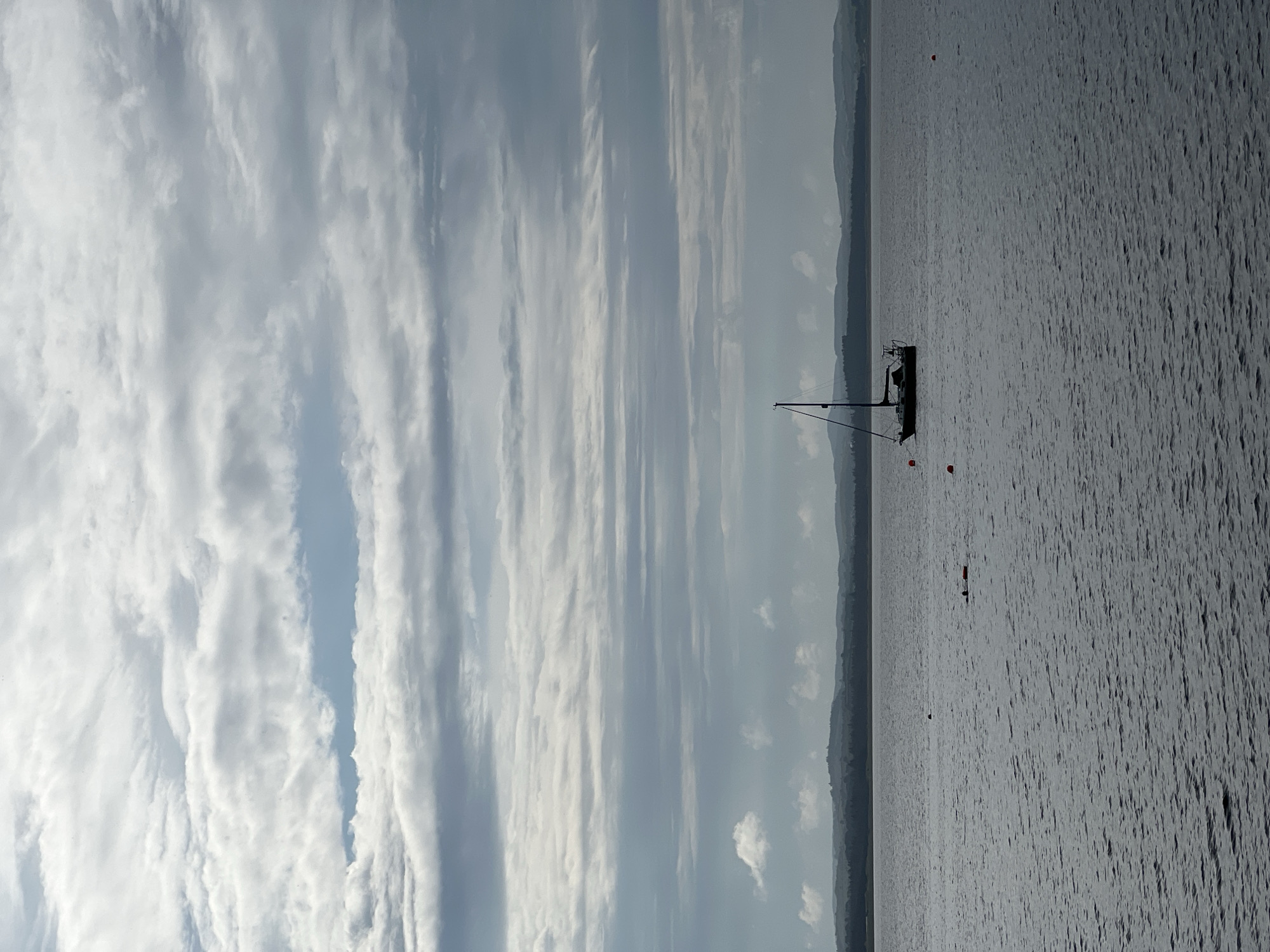 An overcast day in Mountcharles, Ireland, features a calm, grey body of water. A sailboat with a single mast is anchored in the middle distance, silhouetted against the sky. In the foreground, several small, orange buoys are visible. The distant shoreline is lined with dark trees and rolling hills.