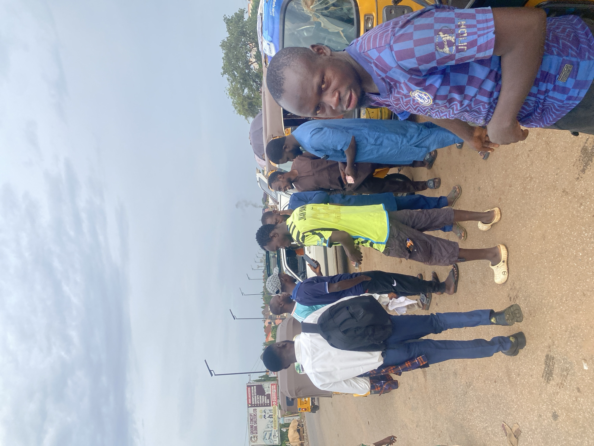 The image depicts a group of about eight men standing along a roadside in Galadimawa Village, Nigeria. The men are diverse in age and attire. One man closest to the camera wears a blue Chelsea jersey. A yellow taxi-like vehicle is nearby. Behind the men is a sign advertising "Ground Concept Global Shelters LTD" with several local locations listed, including Katampe Extension and Karsana. The sky is overcast.