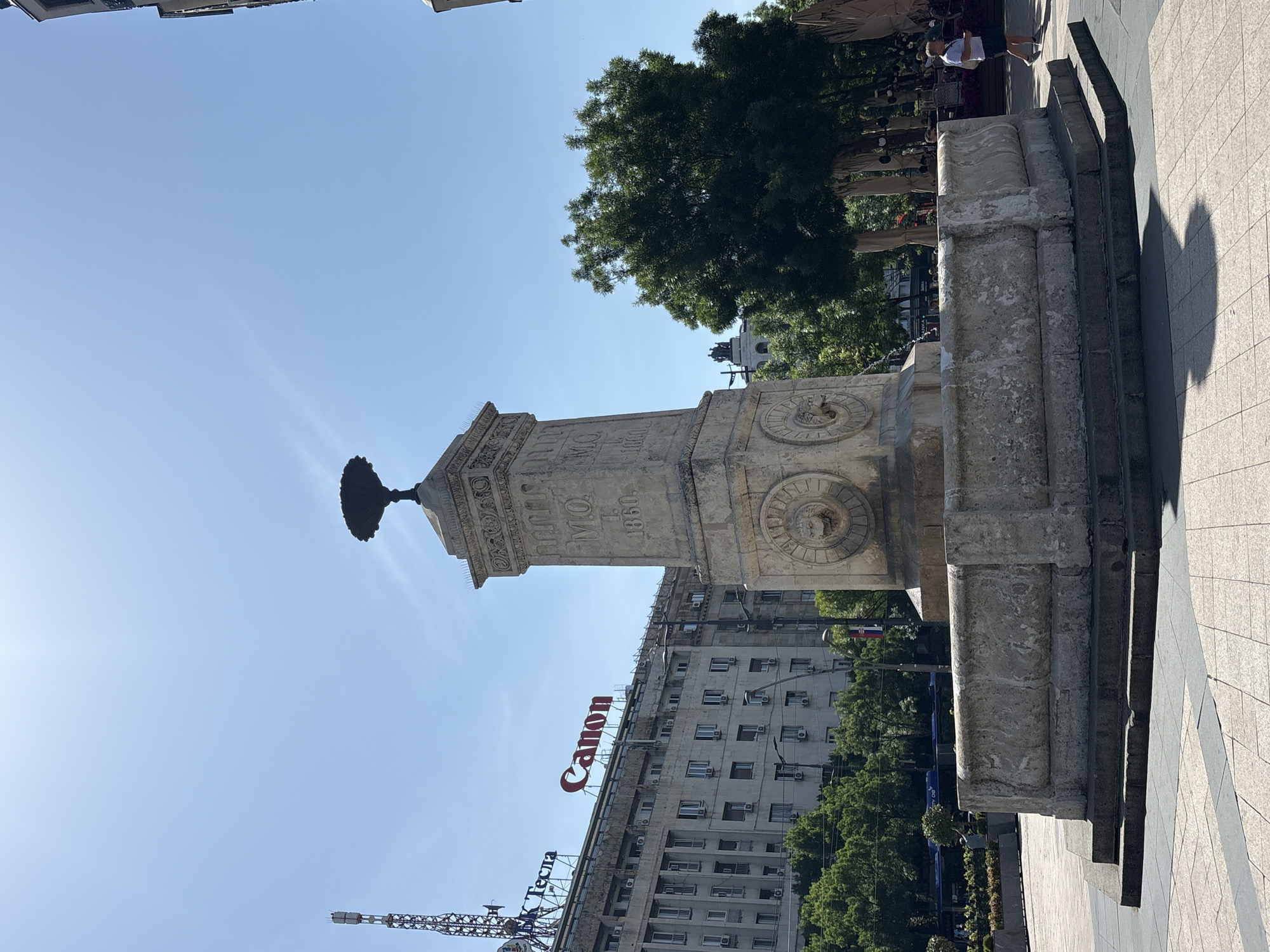 The image shows the Delijska fountain, located in Belgrade, Serbia. The fountain is a stone structure with carved details, including depictions of faces. It is situated on a paved area. In the background, a large building with the word "Canon" in red letters is visible. Further in the background, a cell tower is visible. A tall tree is visible beyond the fountain, and people are present in the distance. The sky is blue.