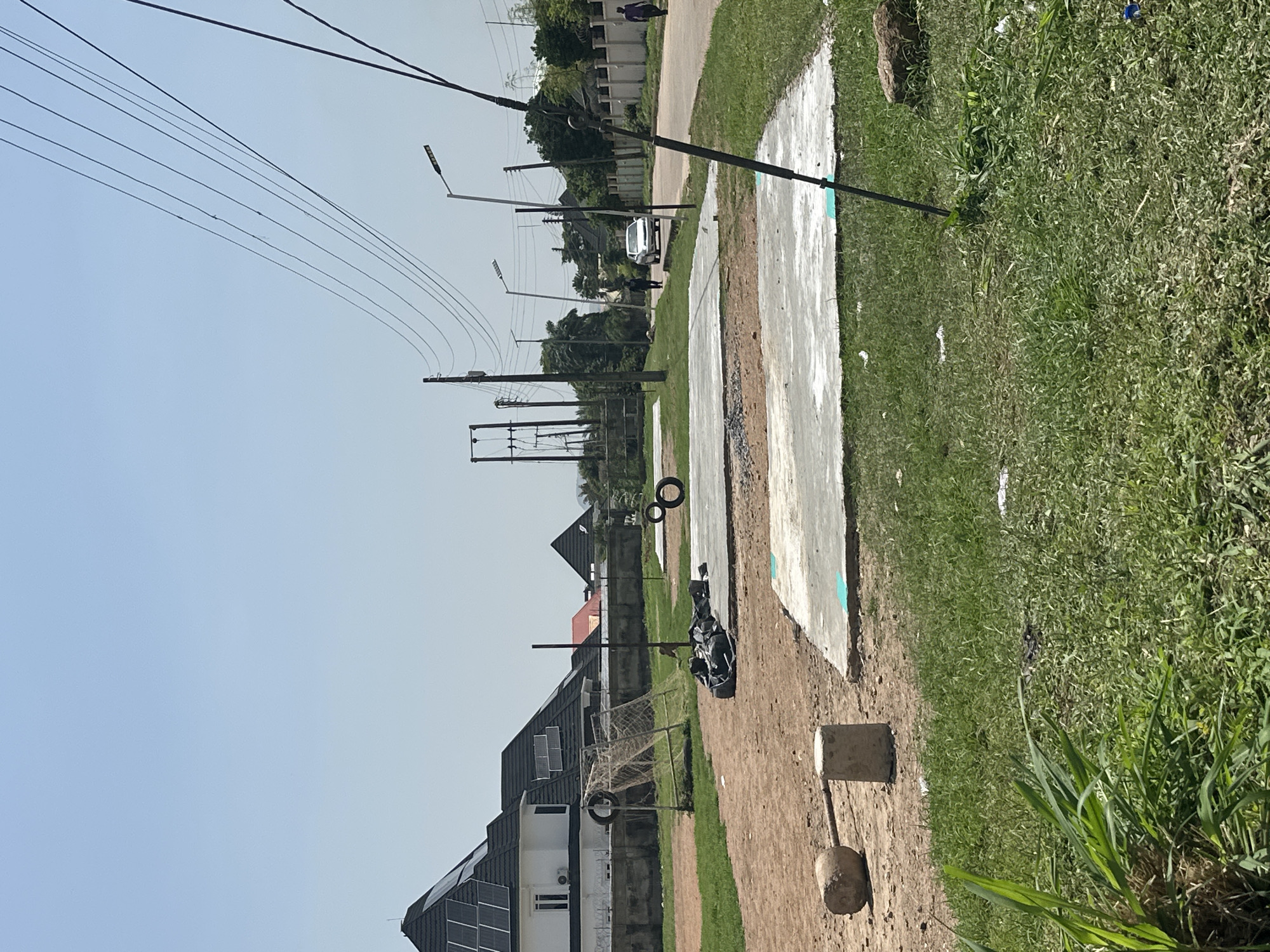 The image depicts an outdoor scene in Abuja, Nigeria, likely a sports or recreational area under development. In the foreground, a patch of green grass is visible, with some scattered debris and a single large rock on the right. A cylindrical concrete marker stands in the dirt on the left, next to a weight made of concrete and metal.
Dominating the mid-ground are two concrete slabs, possibly intended as foundations or playing surfaces, with teal markings on them. Several black tires are scattered around the area, including two placed on one of the slabs and another attached to a dilapidated soccer goal structure on the left. This goal structure has a netting that appears torn. Behind the goal, a modern white house with a dark, pitched roof covered in solar panels is prominent. A concrete wall, topped with barbed wire, encloses this property.
In the background, a street runs from left to right, lined with trees and a white fence. Two streetlights are visible along the street, and power lines stretch across the sky, supported by poles. A silver car is driving away from the camera on the street, and two individuals, one in dark clothing and the other in purple, are walking along the sidewalk. Several other houses and structures are visible in the distance, blending into the urban landscape of Abuja. The sky is clear and blue, indicating a sunny day.
