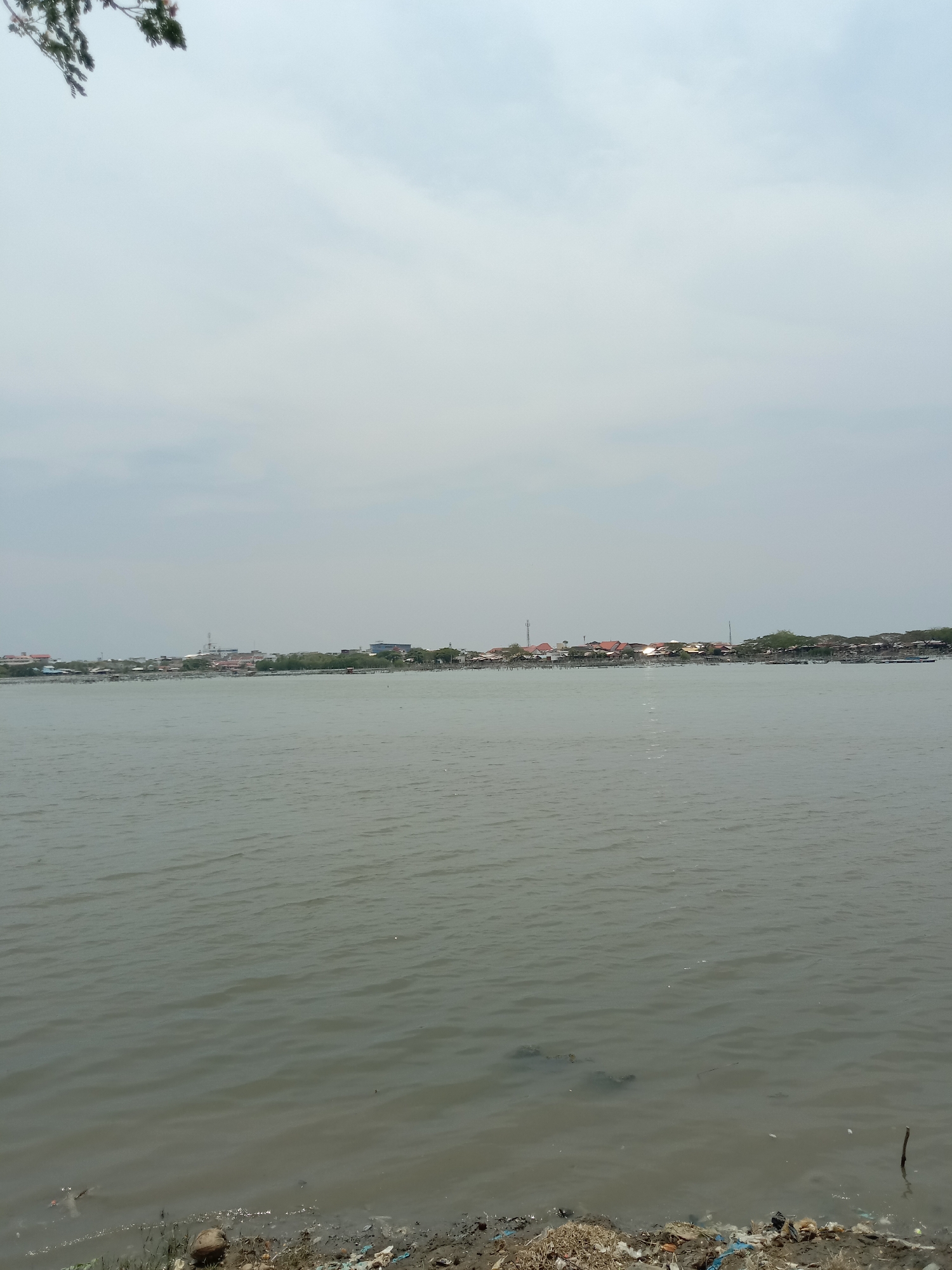 The image depicts a body of water, likely a river or bay, under an overcast sky. The water is slightly choppy with visible ripples. In the distance, a line of buildings and trees indicates a populated area on the opposite bank. The shoreline in the foreground is littered with debris and vegetation. Based on the location information provided (Lhokseumawe, Indonesia), this scene may be a part of a river or coastal area within or near the city. There are no people visible in the image. The environment is characterized by the water, sky, distant shoreline, and littered foreground.