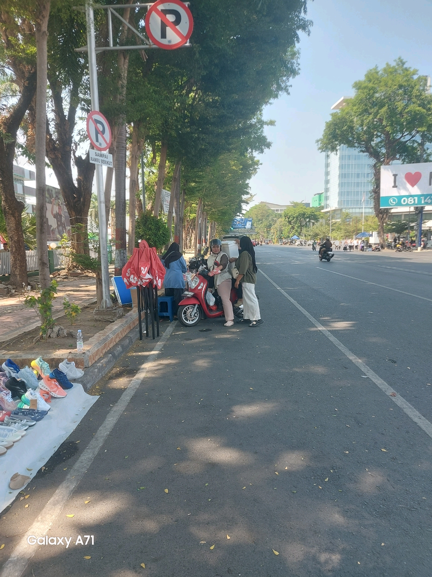 The image depicts a street scene in Makassar, Indonesia, likely during the daytime given the bright sunlight and shadows. In the foreground, on the left side of the road, a vendor is selling shoes displayed on a white tarp. The shoes are varied in color and style, including sneakers and sandals. A plastic bottle of water is also visible near the shoes.
Adjacent to the vendor's stall, a red motorcycle is parked. Three individuals are gathered around the motorcycle, appearing to be interacting with the vendor or each other. One person is wearing a blue top and black headscarf, another is in a light brown top and white pants, and a third is wearing a light brown top with a white headscarf. A red, collapsible umbrella stand is also present, with red umbrellas stacked on it.
Further down the road, traffic can be seen, including several motorcycles and a few cars. A tall, modern glass building stands prominently on the right side of the street, with a billboard featuring the text "I ❤️" and a phone number.
Two "no parking" signs are visible on a pole to the left of the street, with one also indicating "SAMPAI RAMBU BERIKUT" (Until the next sign). Trees line the side of the road, providing some shade. The road itself is paved asphalt with a white line marking the lane. The overall atmosphere suggests a busy urban environment with commercial activity taking place along the roadside.