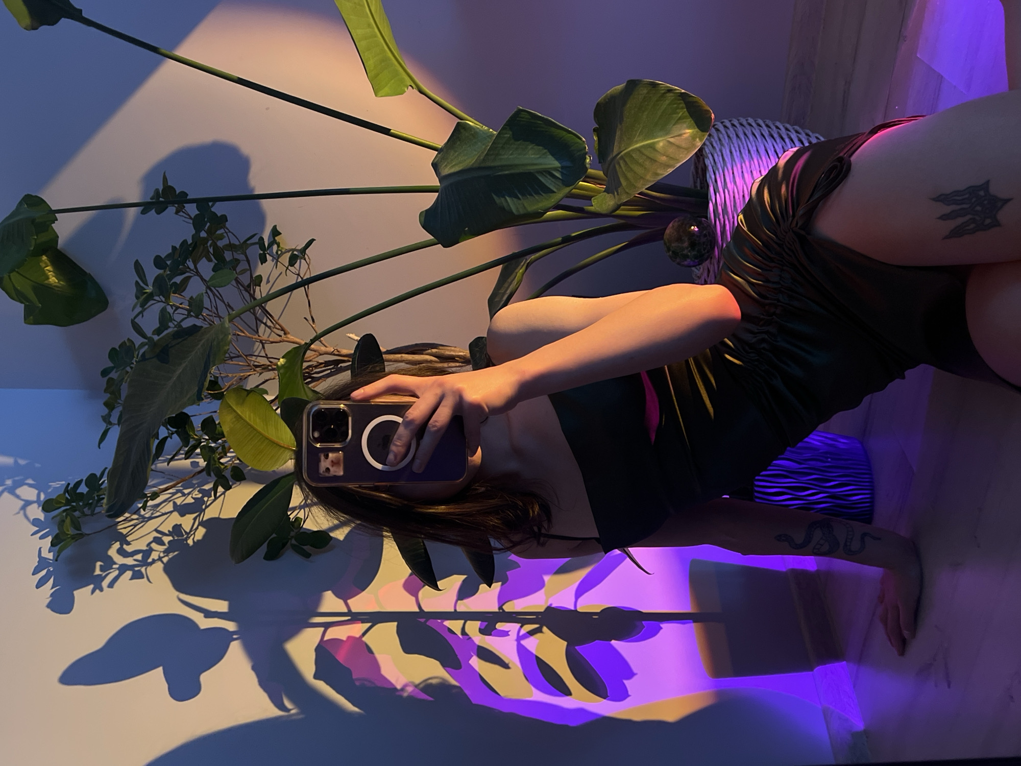 A young woman is taking a selfie in a room with plants and colorful lighting. She is sitting on the floor, leaning against the wall, and holding her phone up to take the picture. She is wearing a black dress and has tattoos on her arm and leg. There are several plants in the room, and the walls are lit with purple and yellow light, casting shadows on the wall. The overall atmosphere is moody and artistic. There are no specific visual cues to determine the location as Ternopil, Ukraine.