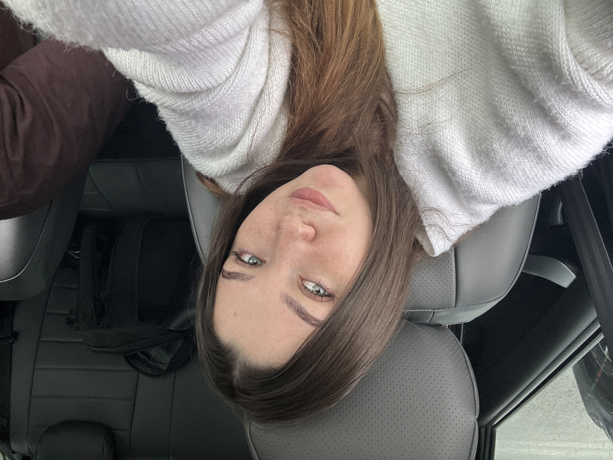 A young woman with long brown hair and striking green eyes is seated in the driver's seat of a car. She is wearing a white knit sweater. The interior of the car appears to be black leather. A dark bag is visible on the back seat. The woman is looking directly at the camera, and her expression is relaxed and approachable. The lighting is natural, suggesting it is daytime. The background is blurred, but the environment appears to be urban. It is not possible to determine the exact location within the city of Perm, Russia from the image alone.