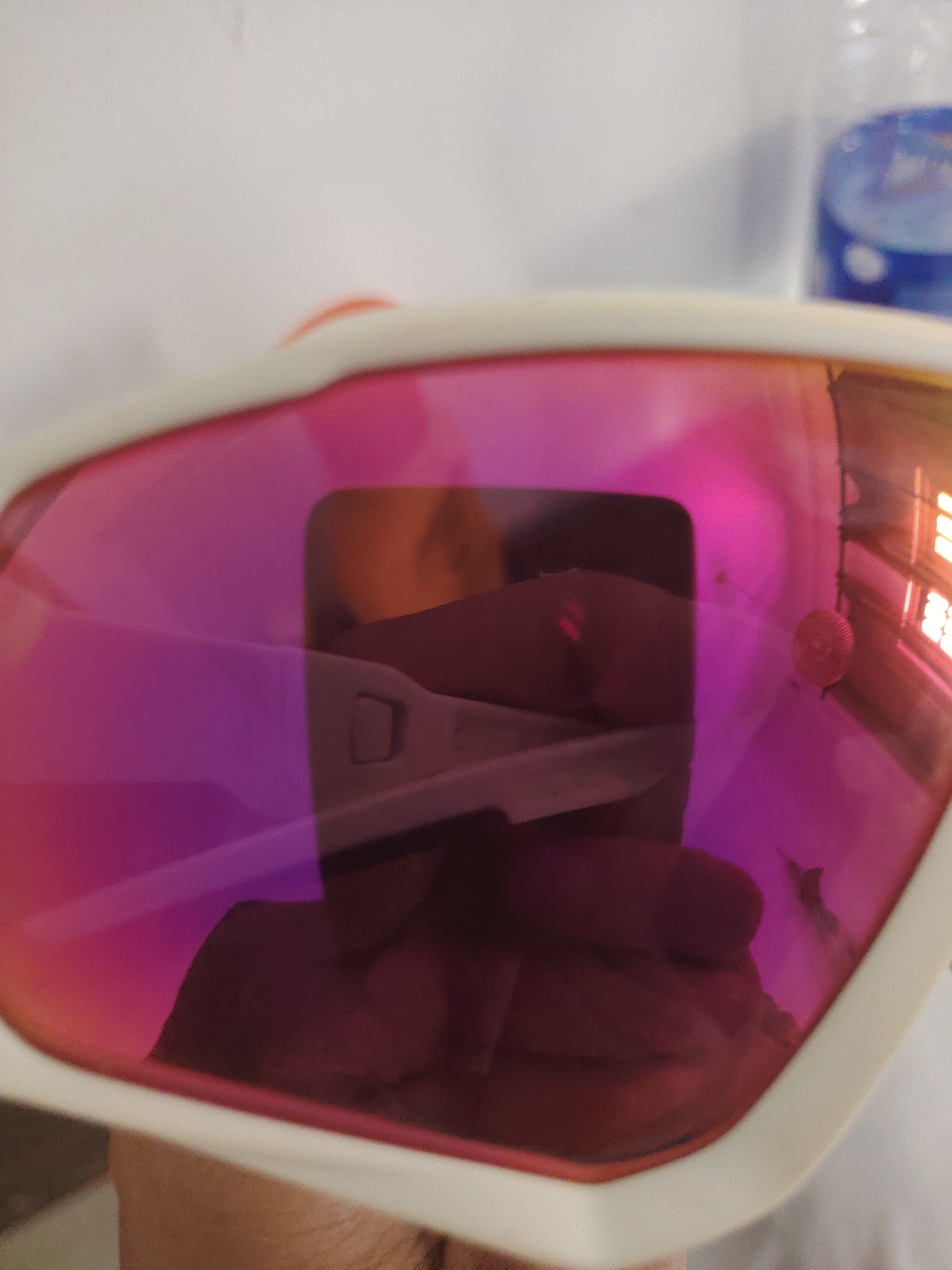 The central focus of the image is a pair of white-framed sports sunglasses with highly reflective lenses. The lenses exhibit a vibrant gradient, shifting from magenta on the left to an orangish hue on the right.
Within the reflective surface of the lenses, a distorted but discernible scene is captured. The most prominent reflection is that of a person's hand, appearing dark and somewhat blurred, holding a white, stick-like object with a small rectangular window. This object strongly resembles a home pregnancy test stick. The fingers gripping the object are clearly visible. Further reflections in the right lens include multiple rectangular shapes, possibly windows or light sources, and a small, spherical, red or pink object, perhaps a decorative item or another light source.
The background of the overall image, outside the sunglasses, is partially visible and largely out of focus. To the top left, a plain, light-colored surface is present. To the top right, a segment of a blue plastic bottle can be seen. The bottom portion of the image shows what appears to be the skin of the hand holding the sunglasses.
The scene implies an indoor setting, illuminated by artificial or indirect light. The primary activity depicted is the act of photographing the reflections within the sunglasses. There is no visible text in the image. The time of day and specific location cues are indeterminate beyond an indoor environment.