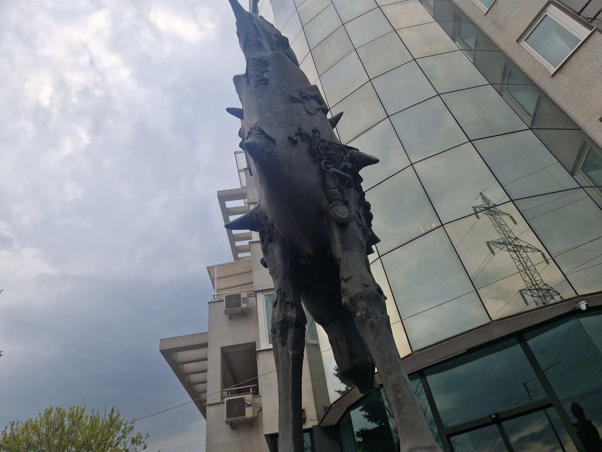 The image captures an urban scene dominated by a large, dark, vertically oriented statue and modern architecture under an overcast sky.
The central subject is a tall, dark-colored statue, possibly made of bronze or dark stone, occupying the mid-ground. It has a rough, textured surface with various abstract or stylized features, including spikes or protrusions along its length and intricate carvings, particularly visible on its lower section. The statue stands upright, appearing to be on two thick, irregular supports, viewed from a low angle, emphasizing its height and imposing presence.
To the right of the statue is a modern building with a curved, highly reflective glass facade. The glass panels show a clear reflection of a large electricity transmission tower (pylon), providing a significant environmental detail. Above this, the building's upper floors are visible, constructed from a lighter-colored material, featuring rectangular windows.
To the left and behind the statue, another building with a lighter, perhaps concrete or stucco, exterior is visible. This building has multiple balconies, some with railings, and several visible air conditioning units attached to its walls.
The sky is overcast with a mix of light and darker grey clouds, suggesting a cloudy daytime. A small patch of green foliage from a tree is visible in the bottom left corner, grounding the urban elements with a touch of nature. No specific activity is taking place, and there is no visible text. The overall scene presents a blend of art and architecture in a city environment.