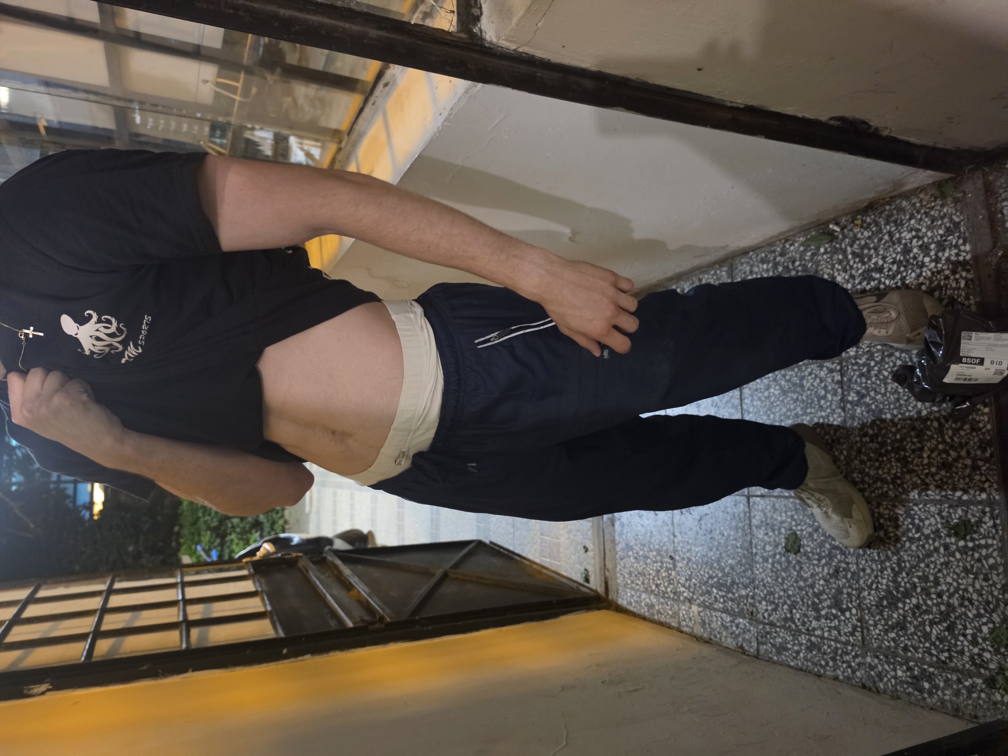 A person stands in a narrow, open-air walkway between two buildings, possibly an alleyway or side entrance. The walkway is paved with speckled grey tiles and has some green debris scattered on the ground. On the left, a wall painted yellow transitions to a cream-colored, textured wall. Adjacent to this wall is a dark metal gate with vertical bars. On the right, a cream-colored wall with a similar textured finish is visible, bordered by a dark metal frame.
The person is wearing a black t-shirt with a white octopus logo and the text "MC SPORTS" underneath, a white waistband peeking out from their light-colored briefs, dark blue track pants with white piping detail, and light tan athletic shoes. The person is lifting the hem of their t-shirt, revealing their abdomen and a thin, dark scar running vertically across their stomach. Their right hand is near their left pocket. In the foreground on the right, a black plastic package with white labels is on the ground. The lighting suggests it is either dusk or nighttime, with artificial light sources casting shadows. The scene does not contain any specific visual cues that place it definitively in Sofia, Bulgaria.