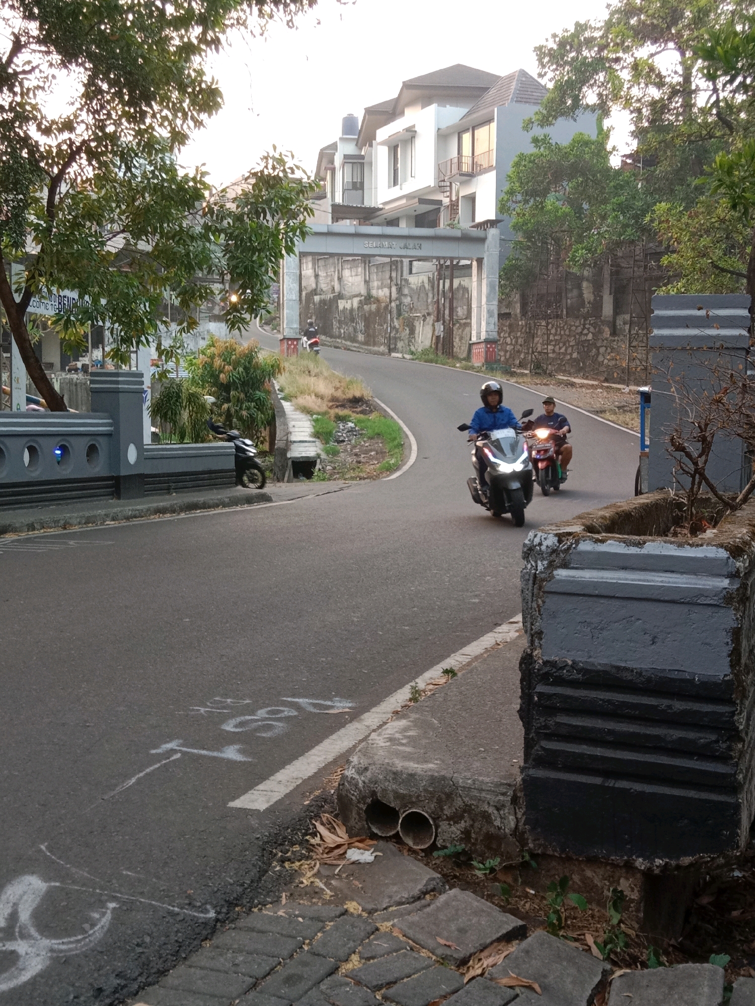 The image depicts a road winding uphill in a residential area, possibly within the city of South Tangerang, Indonesia, given the presence of modern multi-story houses and a gate with what appears to be Indonesian text. Two individuals are riding motorcycles on the road. The first motorcycle, a scooter, is closer to the foreground, ridden by a person wearing a blue jacket and helmet. Behind them, another person on a motorcycle, wearing a dark shirt and cap, is also visible. The road is paved and has a white line marking its edge. The surroundings include trees, vegetation, and concrete structures, including a gray, ornate fence on the left and a darker, textured concrete planter on the right. Some white markings are present on the road surface. The time of day appears to be late afternoon or early morning, with soft lighting.
