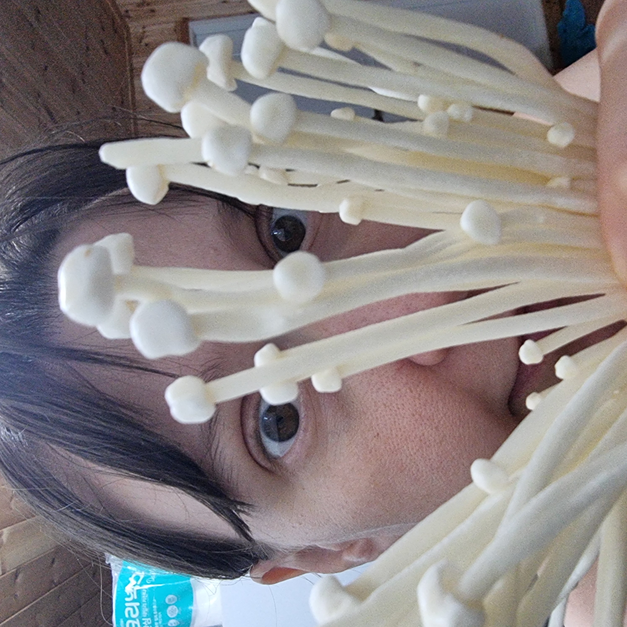 A close-up shot shows a person's face, partially obscured by a cluster of white enoki mushrooms. The person, whose skin appears light-toned, has dark hair that falls around their face, and their eyes are wide and looking towards the camera. The mushrooms are thin and elongated, with small white caps. The background shows a wooden-paneled wall and a portion of a blue and white package. The scene appears to be indoors.