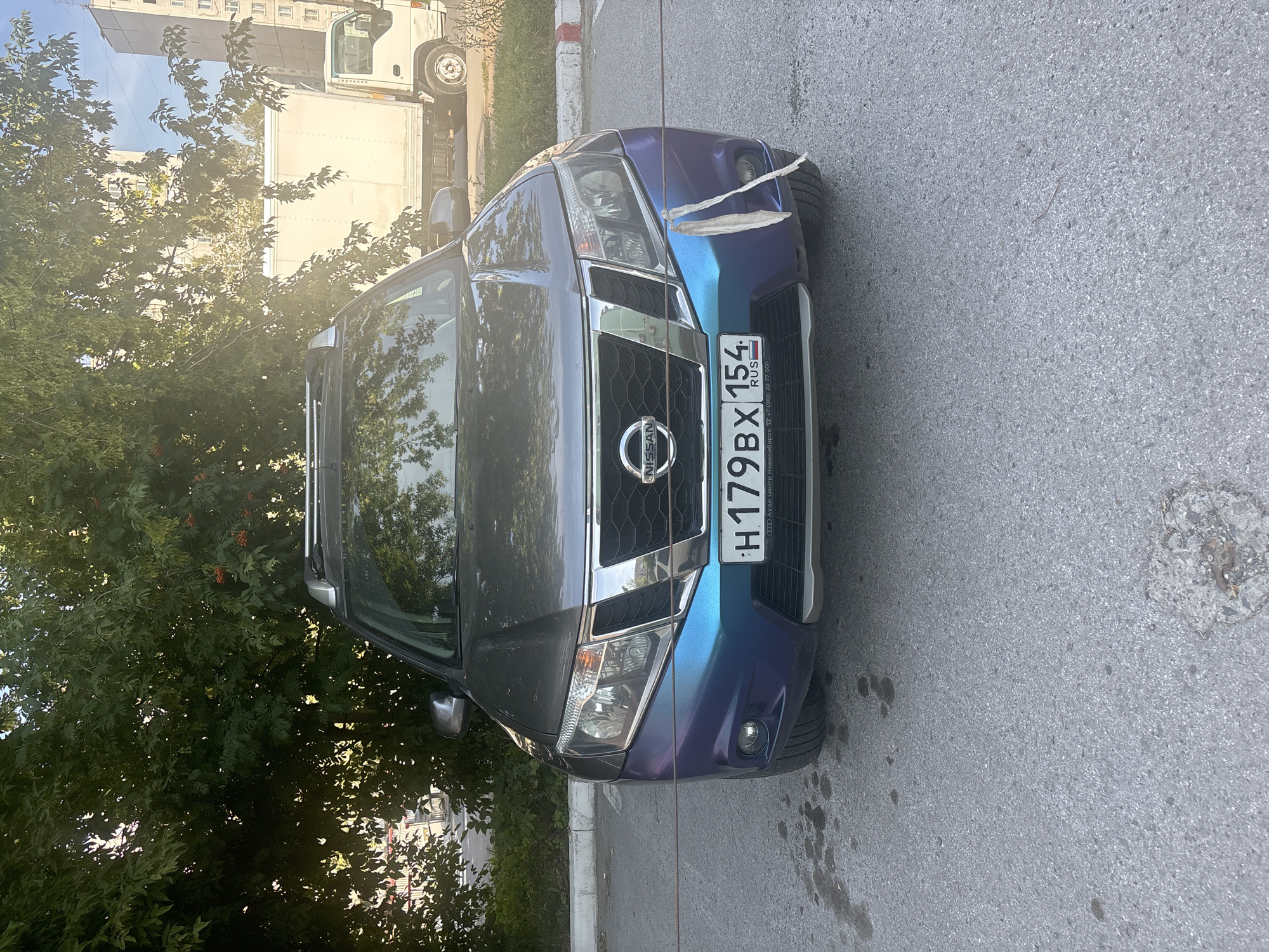 A Nissan SUV is parked on an asphalt lot in front of a row of trees. The car's front bumper has a blue and purple gradient paint job. A white piece of tape is hanging from the right side of the bumper. The car's license plate reads "H179BX154 RUS". The bottom of the license plate shows the text "ООО Ауди Центр Новосибирск" and a phone number. In the background, a white box truck is parked and a modern apartment building is visible. The scene is well-lit, suggesting it is daytime.