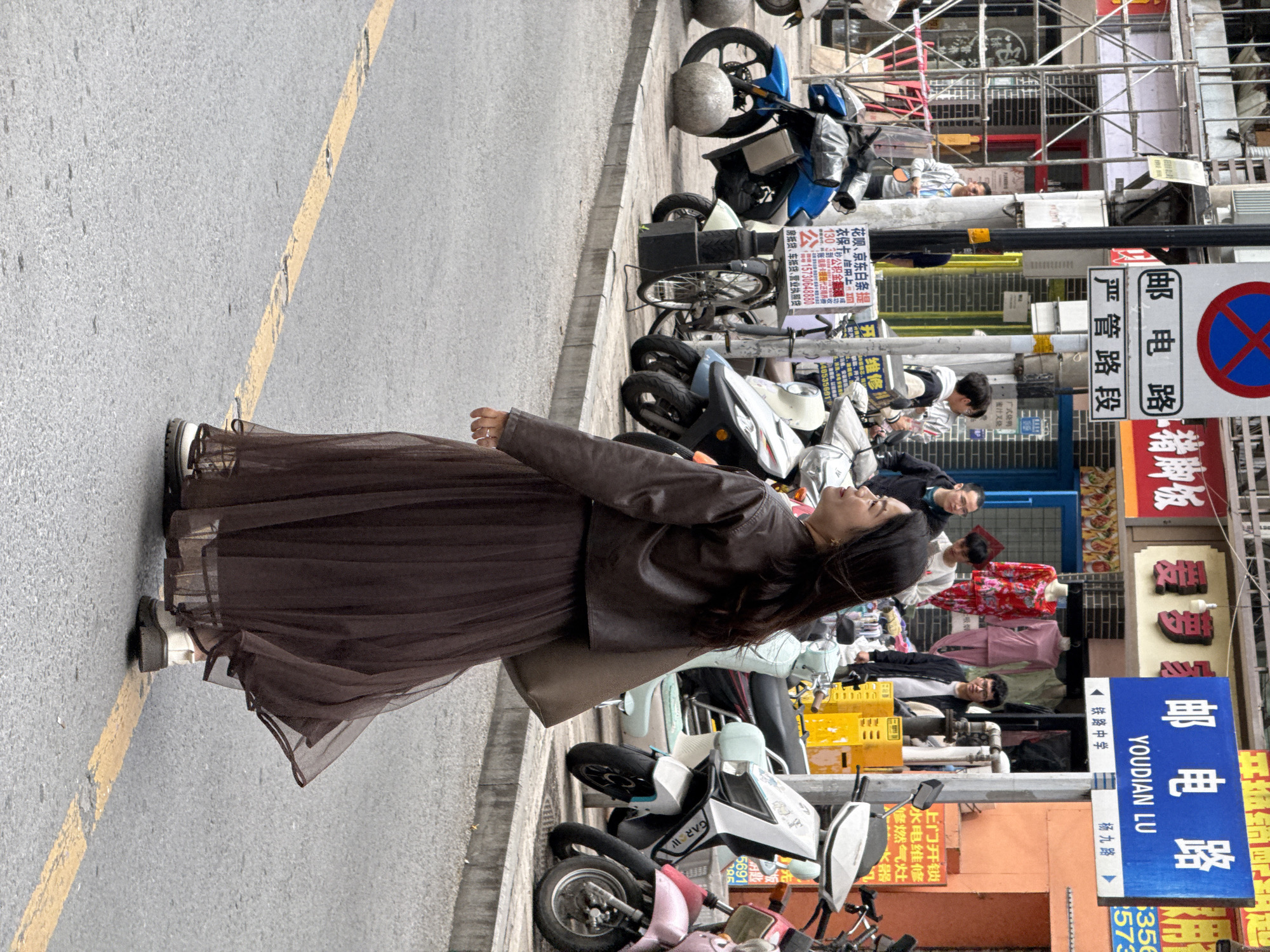 The image displays a street scene on Youdian Lu in Jiulongpo District, China. A woman is captured from behind, looking toward her left, walking on an asphalt road marked with a faded yellow line. She has long dark hair and is dressed in a dark brown cropped jacket over a long, flowing dark brown sheer skirt, paired with light-colored, thick-soled shoes. A light brown shoulder bag is carried on her right side.
To her right, a raised concrete sidewalk hosts a dense arrangement of parked motorcycles and electric scooters, including models in blue, light green, white (branded "GARII"), and pink. Several individuals, primarily men, are visible on the sidewalk in the mid-ground, some standing near commercial storefronts. The background consists of multi-story commercial buildings featuring various Chinese signage, such as "猪脚饭" (Pig's Feet Rice) and "爱罗," with one shop displaying mannequins dressed in red and pink loungewear. Construction scaffolding is present on a building further down the street on the left.
Prominent street furniture includes multiple utility poles, one bearing a blue rectangular sign identifying the street as "YOUDIAN LU" with directional arrows for "铁路中学" (Railway Middle School) and "杨九路" (Yangjiu Road). Another pole displays a red 'X' in a blue circle "No Parking" sign mounted above a sign stating "邮电路 严管路段" (Youdian Lu, Strictly Managed Road Section). Additional Chinese advertisements for services such as "花呗、京东白条提衣保卡、座垫" (Huabei, Jingdong Baitiao, clothing insurance card, seat cushion) and "维修改装" (Repair and Modification) are affixed to the poles. Two grey spherical concrete bollards are situated on the sidewalk near the blue motorcycle.