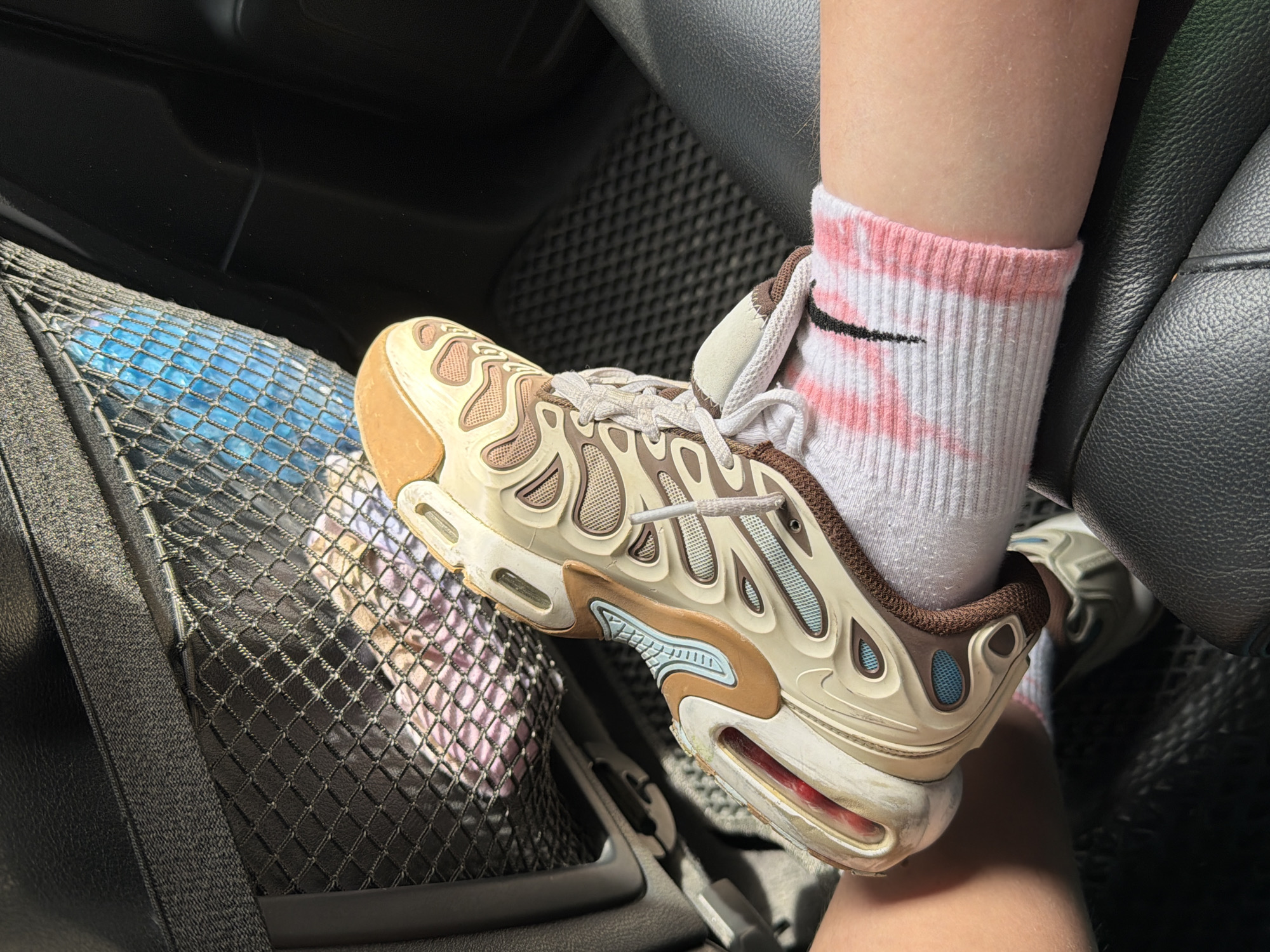 The image shows a close-up view of a person's foot wearing a white sock with pink stripes and a beige and brown sneaker. The foot is resting on a black car seat. In the background, a mesh organizer is attached to the back of a car seat, containing several items, including what appears to be blue water bottles and pink fabric. The lighting suggests it is daytime, with sunlight casting shadows. The overall scene is indoors, within a vehicle. The provided location of Катунь, Russia is external to the visual information present in the image.
