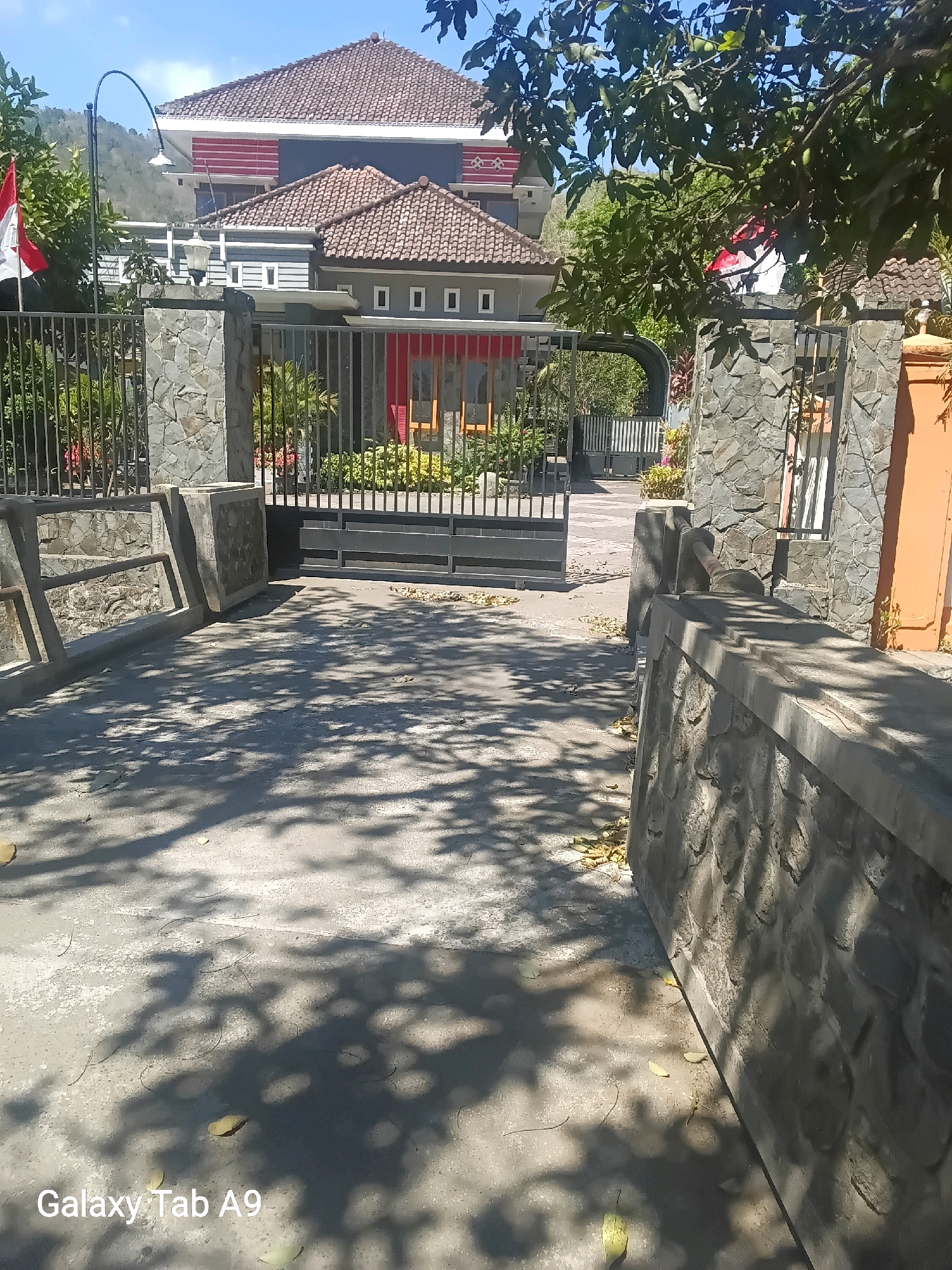 The image depicts the entrance to a property in Trenggalek, Indonesia, characterized by a stone-clad fence and gate. A large, modern house with a red and white striped section on its upper facade and terracotta tiled roofs is visible behind the gate. The house has multiple windows and is surrounded by lush green foliage. The gate itself is made of dark metal bars and appears to be closed. In the foreground, a concrete pathway is visible, partially covered by shadows cast by trees. A lamppost with a curved arm stands to the left of the gate. There are no people present in the image, and the scene appears calm and unoccupied. The overall environment suggests a residential area with well-maintained landscaping.