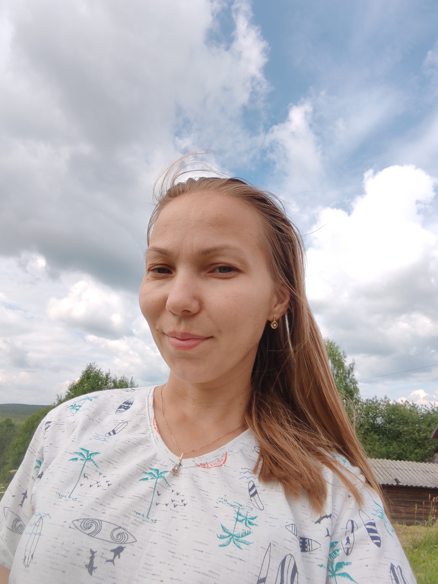 A fair-skinned woman with long, light brown hair stands outdoors, taking a selfie. She is wearing a white t-shirt with a repeating pattern of surfboards, palm trees, and small birds. A delicate gold chain with a pendant is visible around her neck. She has on a small, square gold earring with a clear stone in her right ear. Her expression is relaxed, with a slight smile.
The background consists of a cloudy sky with patches of blue peeking through. Below the sky, a green, rolling landscape is visible with trees and distant hills. To the right of the woman, a rustic wooden building with a grey corrugated roof can be seen. The overall impression is of a casual outdoor setting, possibly in a rural area outside of Чухлэм, Russia. The lighting suggests it is daytime, with diffused light from the overcast sky.