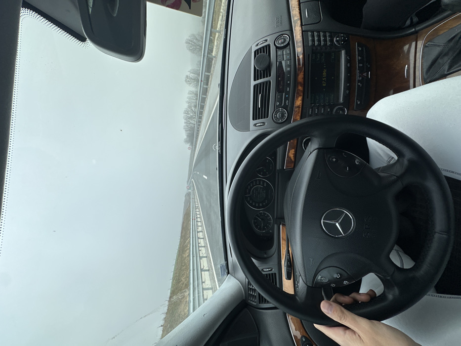 The image captures a driver's-eye view from inside a Mercedes-Benz vehicle, likely an E-Class, traveling on a highway near Cherencha, Bulgaria, under heavily overcast or foggy conditions. A person's light-skinned left hand is positioned on the black leather steering wheel, which features the Mercedes-Benz logo and "SRS AIRBAG" in its center. The dashboard incorporates black materials and wood grain trim. The instrument cluster displays a speedometer needle indicating approximately 100-110 units (likely km/h), alongside other gauges. The center console features an audio unit displaying "87.5 MHz" and climate controls, with a circular black magnetic phone mount affixed above the central vents. Through the windshield, a multi-lane highway is visible, stretching into a hazy distance under a uniformly gray sky. Metal guardrails line both sides of the road. An earthen embankment with sparse brown vegetation is present on the left, while on the right, bare deciduous trees extend into the fog. A red circular speed limit sign is faintly discernible in the distance on the right side of the highway. The road surface appears to be asphalt, possibly damp.