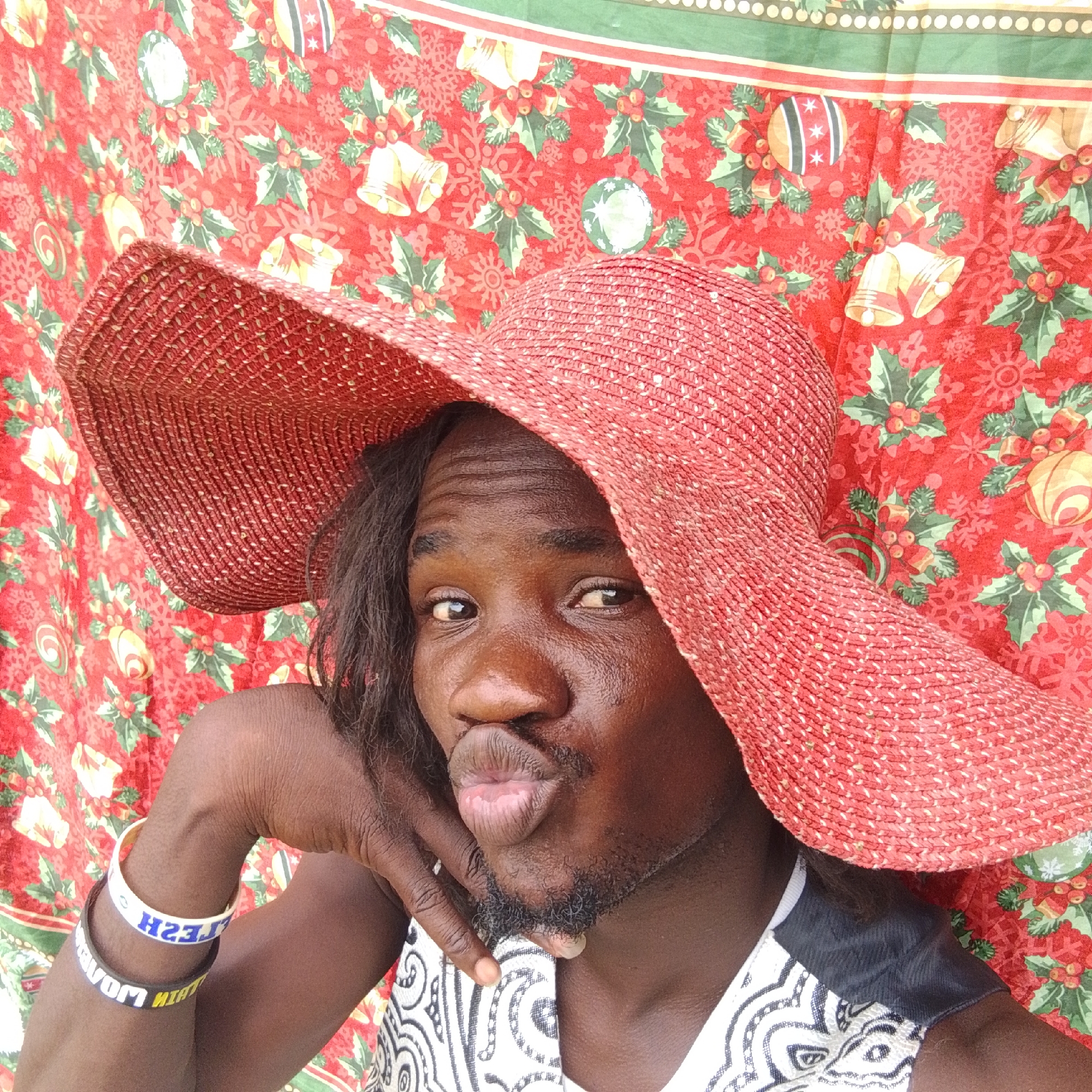 A person is leaning their head on their hand, wearing a red wide-brimmed hat and a patterned white and black top. They are posing for a selfie against a red backdrop with a Christmas-themed pattern. Two bracelets are visible on their arm. The image appears to be taken outdoors, possibly in a residential area, given the presence of fabric as a backdrop. There are no specific landmarks or architectural features visible that would allow for precise location identification within Ado, Nigeria. The scene is static, with the subject posing.