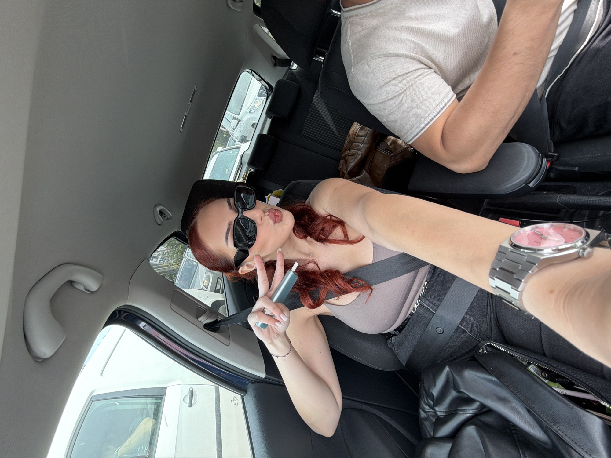 The image shows a selfie taken inside a car. A young woman with red hair is in the driver's seat, wearing sunglasses, a taupe top, and a seatbelt. She holds a vape pen and makes a peace sign with her fingers. She has a watch on her wrist. There is a passenger to her right. Other vehicles are visible outside the window. Based on the visible cars, the location could plausibly be in Sofia, Bulgaria.