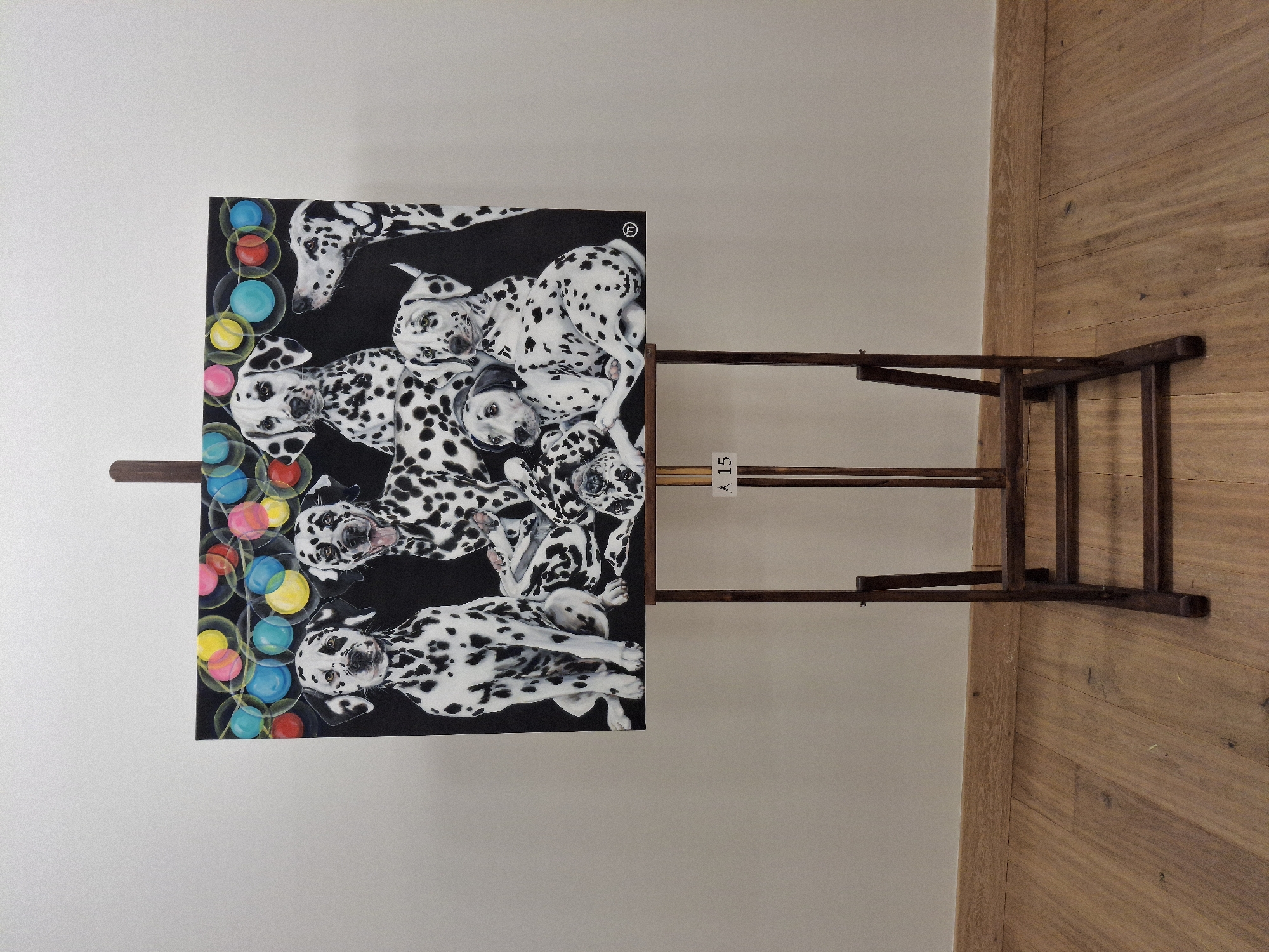 An indoor scene features a vibrant painting, rotated 90 degrees clockwise from its intended horizontal orientation, displayed on a dark wooden easel. The painting depicts several playful Dalmatian dogs with their characteristic black spots on white fur, set against a black background. The dogs are captured in various poses, some looking directly out, others appearing to be resting or engaged in activity. Along the left edge of the artwork, a vertical column of colorful, translucent bubbles in red, blue, yellow, pink, and green adds a whimsical element. A small, circular logo, possibly an encircled 'P' or similar, is visible in the top right corner of the painting.
The easel itself is made of dark wood, providing a sturdy support for the artwork. A small white tag with the black text "A 15" is attached to one of the easel's horizontal bars. The background consists of a plain, light-colored wall, likely off-white or light gray, and to the right, a light brown wooden floor with visible planks. The overall setting is simple, focusing attention on the artwork.