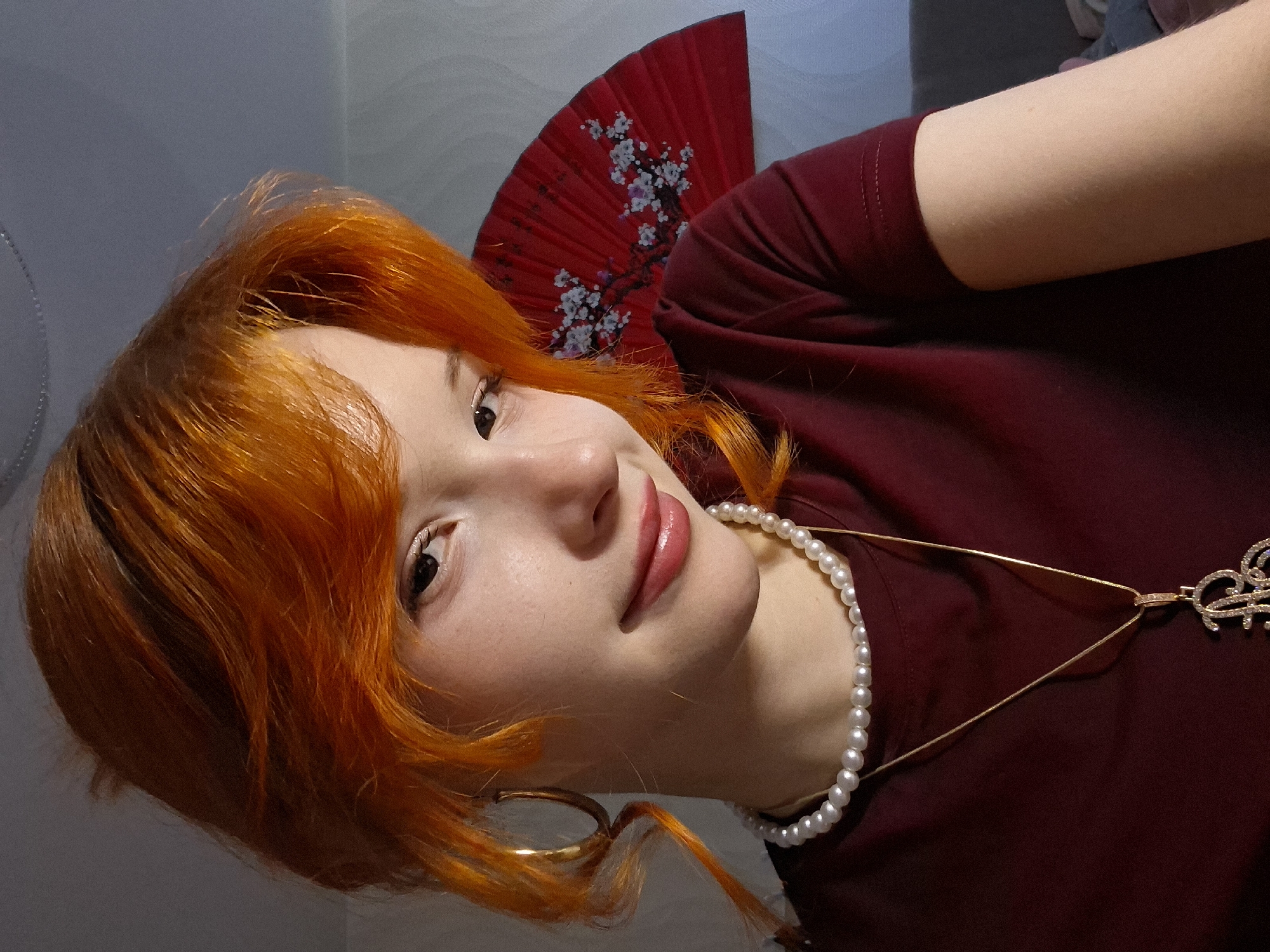 A portrait of a young woman with vibrant orange hair is presented from a close-up, tilted perspective. She is wearing a maroon top, a pearl necklace, and a gold necklace with a decorative pendant. One gold hoop earring is visible. A red fan with a white floral design is positioned behind her head. The background appears to be a neutral-toned wall. There is no information to conclude that the image was taken in the city of Chaikovsky, Russia.